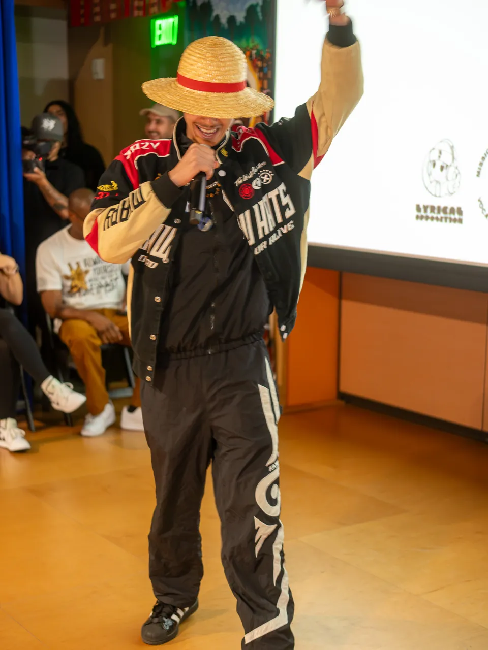 A performer in a straw hat raises an arm while holding a microphone beside the screen.