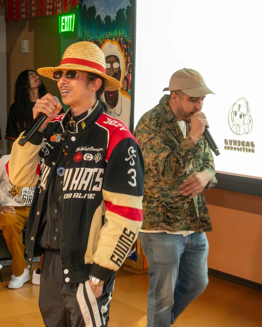 Two performers share the floor with microphones, one in a patterned jacket and one in a straw hat and racing jacket.