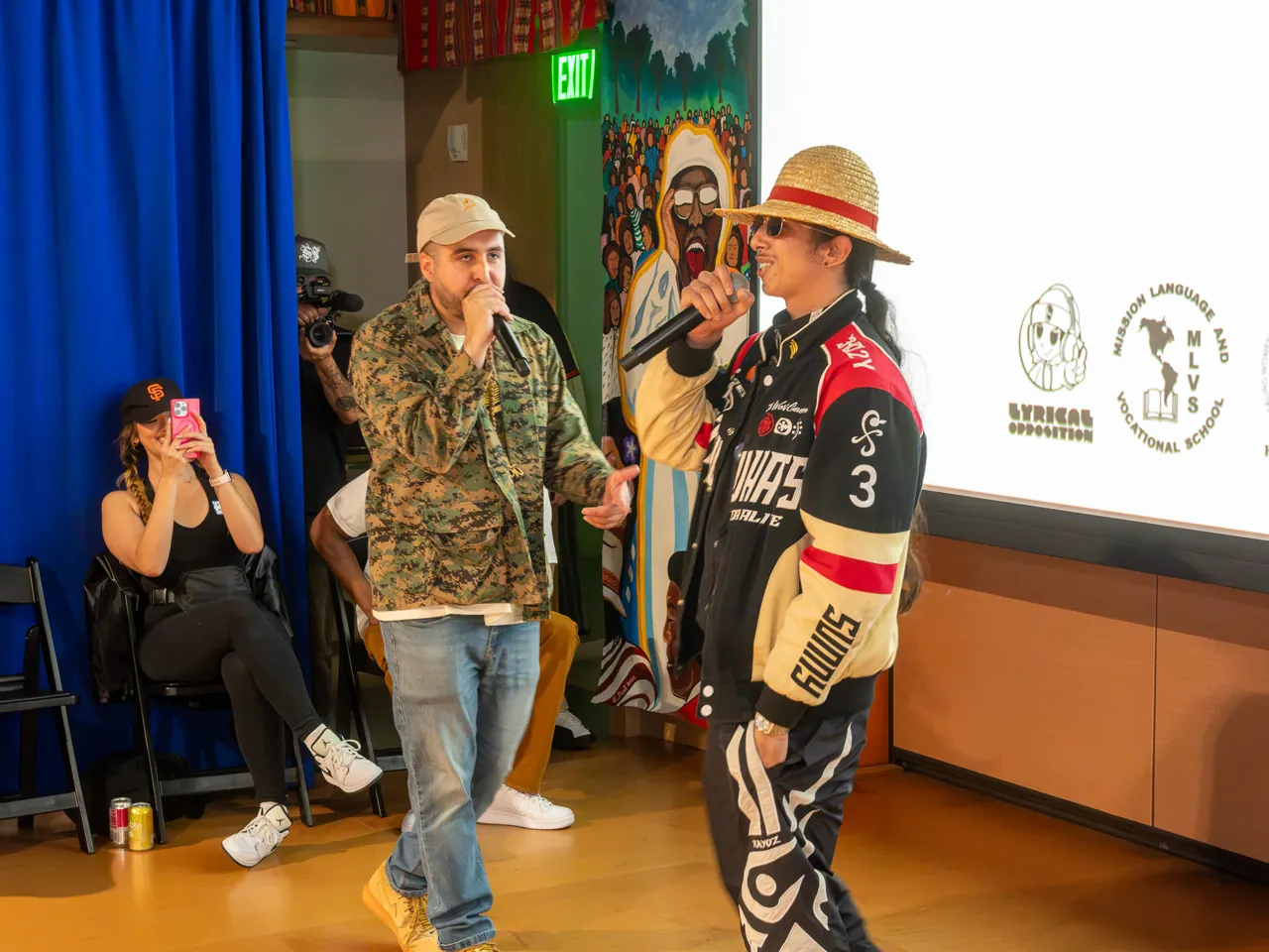 Two performers share the floor with microphones, one in a patterned jacket and one in a straw hat and racing jacket.