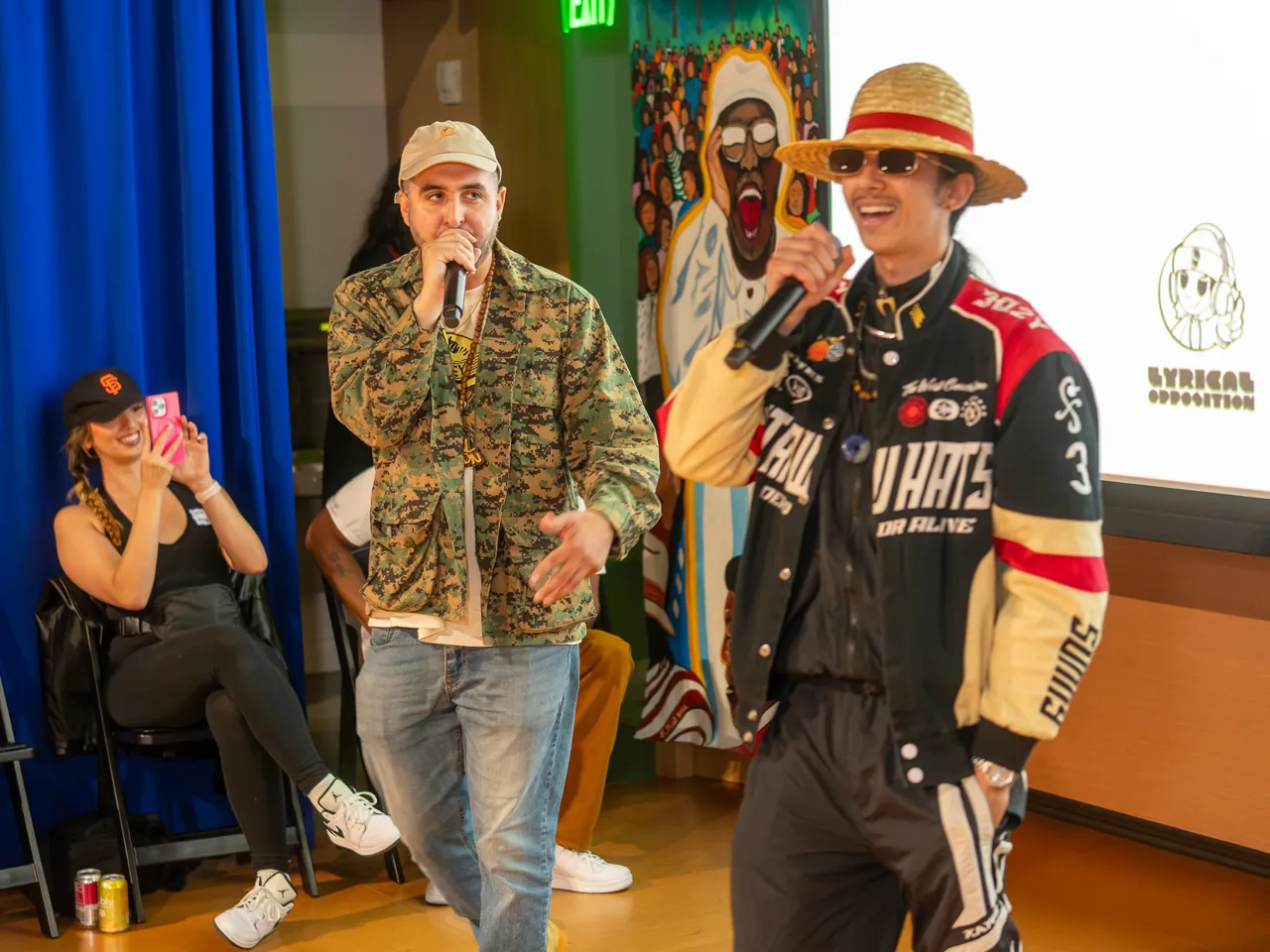 Two performers share the floor with microphones, one in a patterned jacket and one in a straw hat and racing jacket.