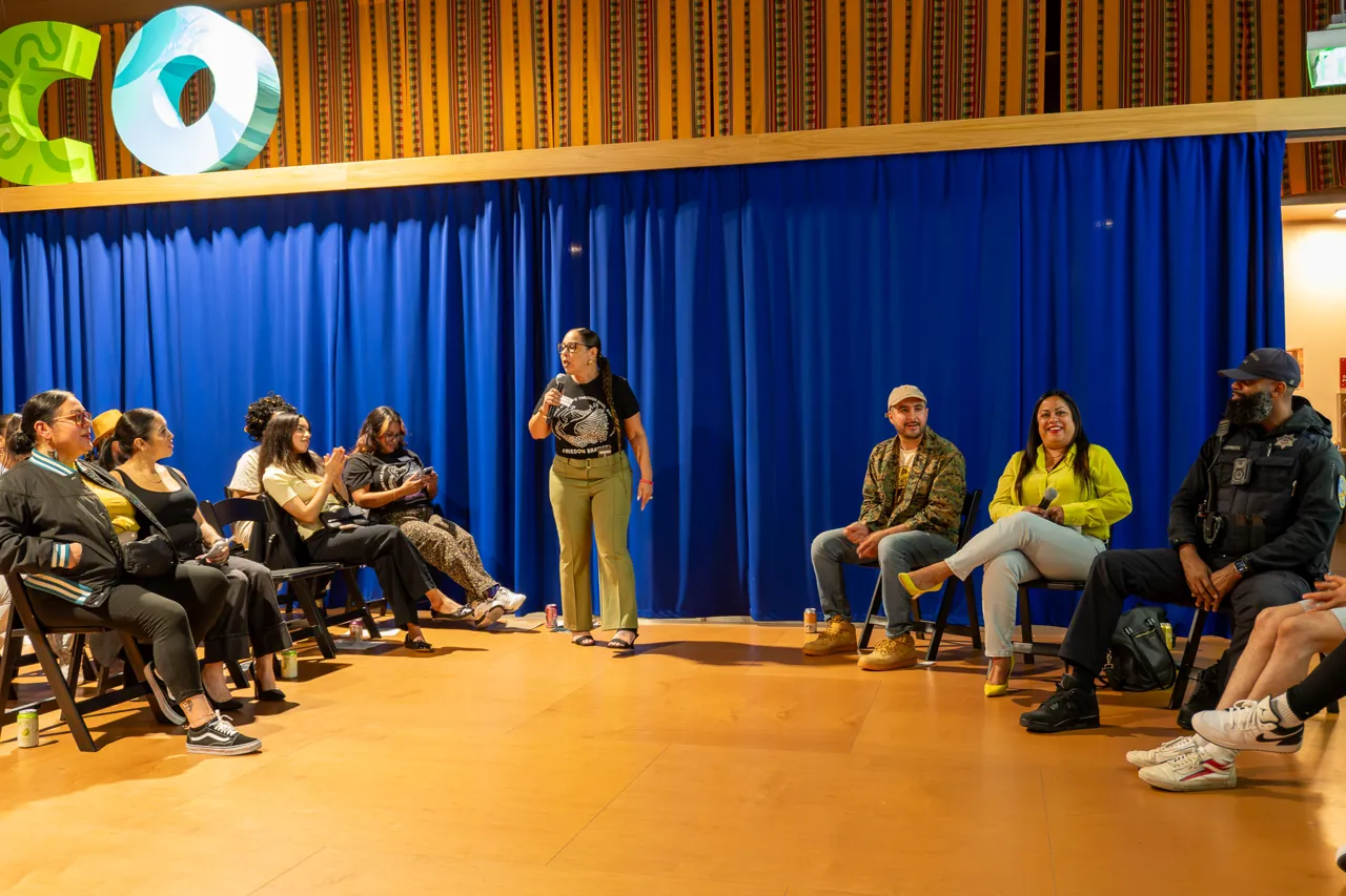 A speaker stands in the open floor area between the seated audience and panel.
