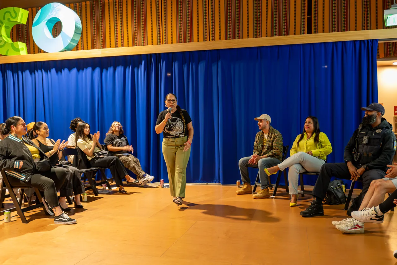 A speaker stands in the open floor area between the seated audience and panel.