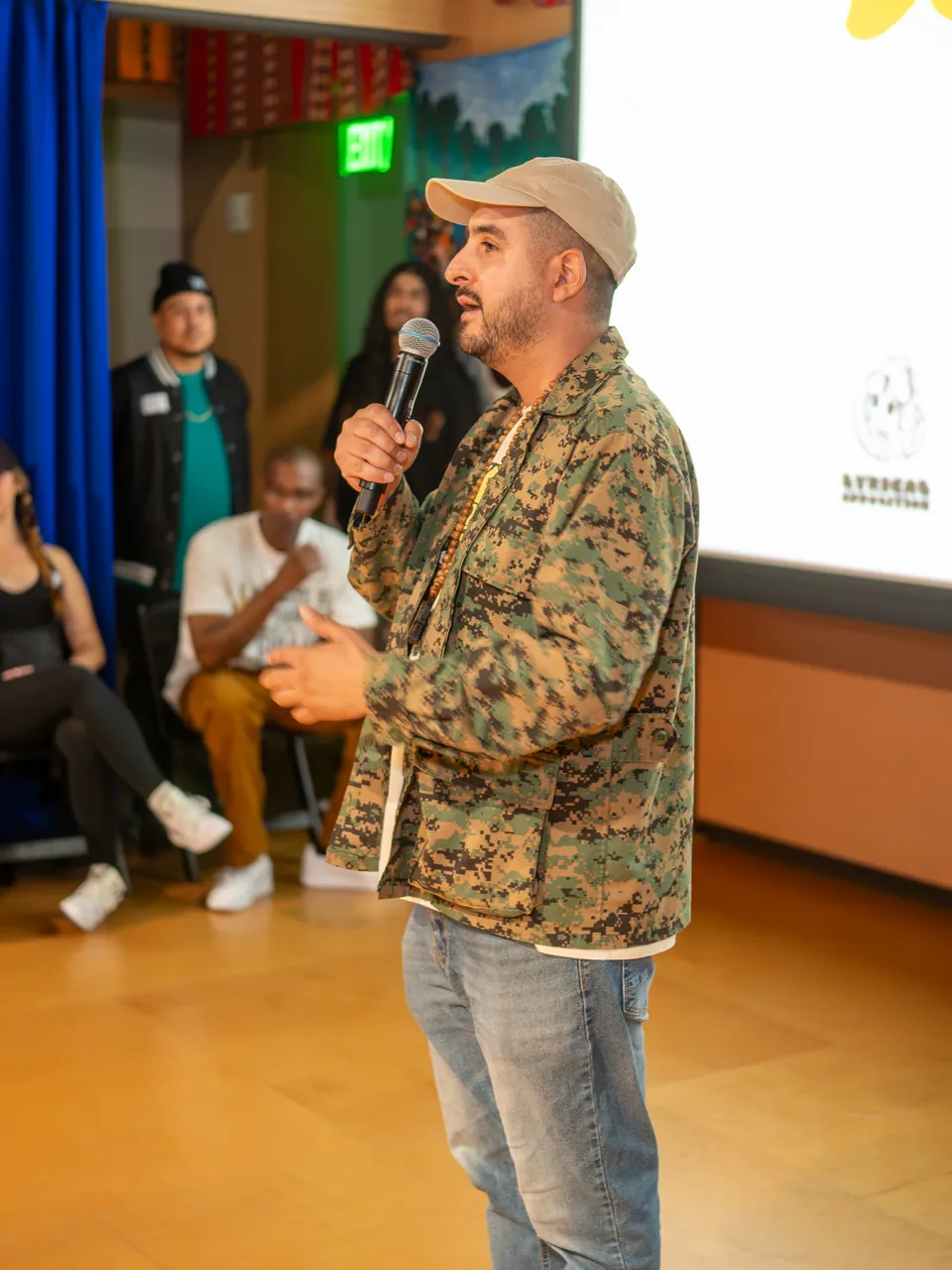 A speaker in a cap and patterned jacket addresses the room with a microphone.