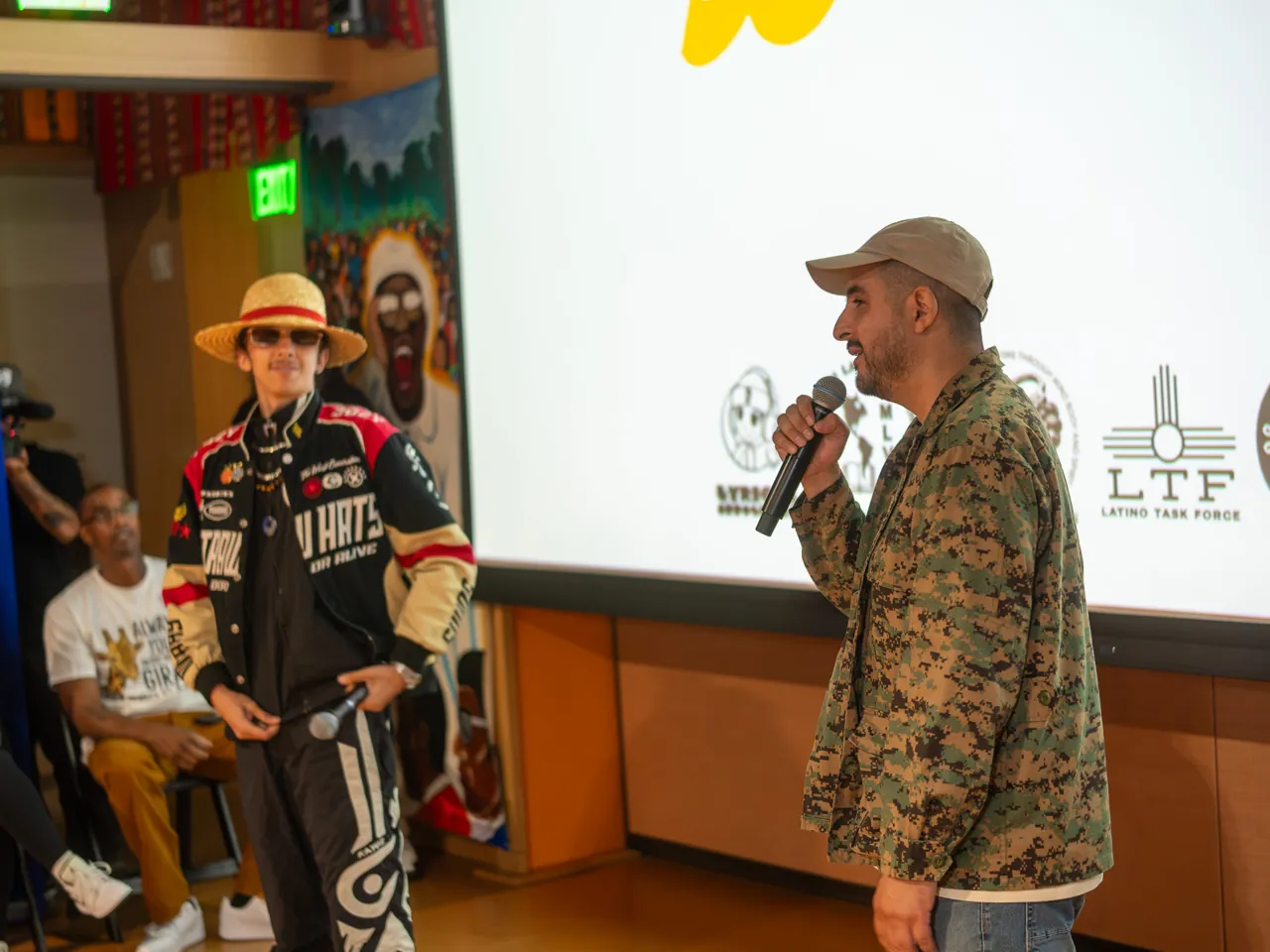 Two performers share the floor with microphones, one in a patterned jacket and one in a straw hat and racing jacket.
