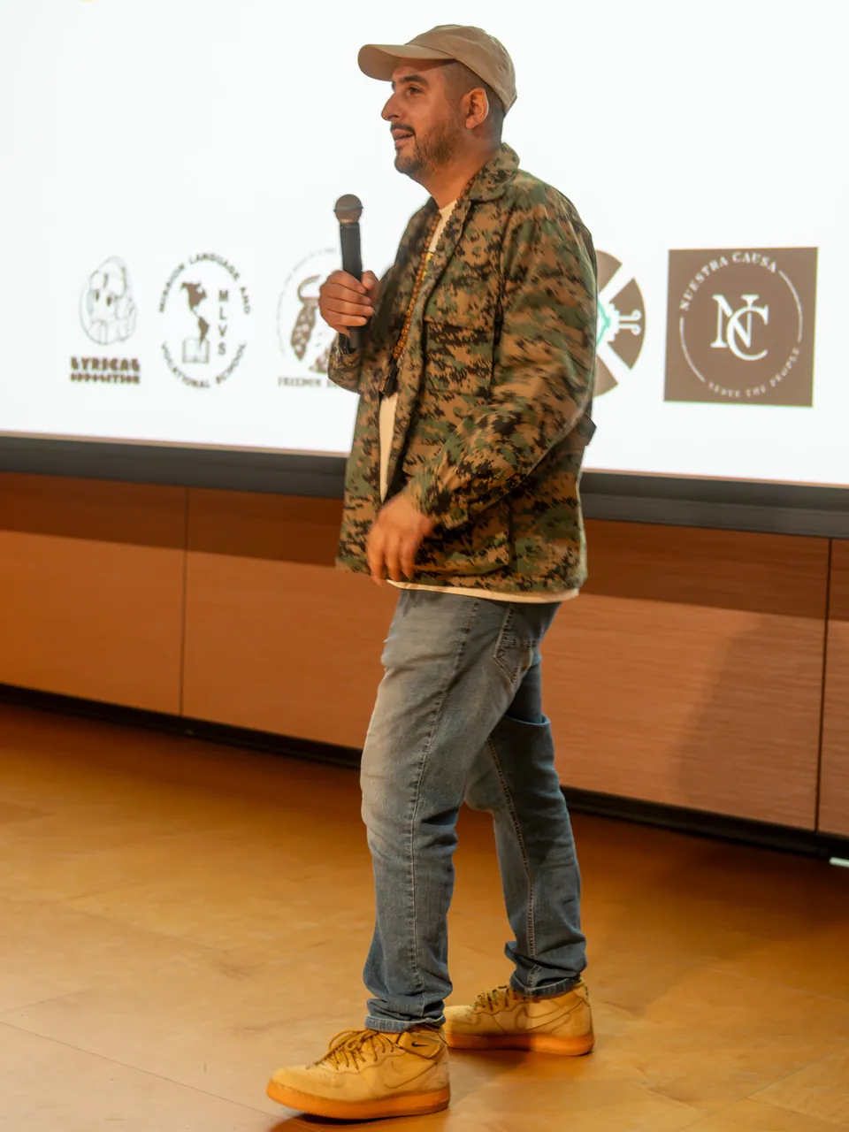 A speaker in a cap and patterned jacket addresses the room with a microphone.