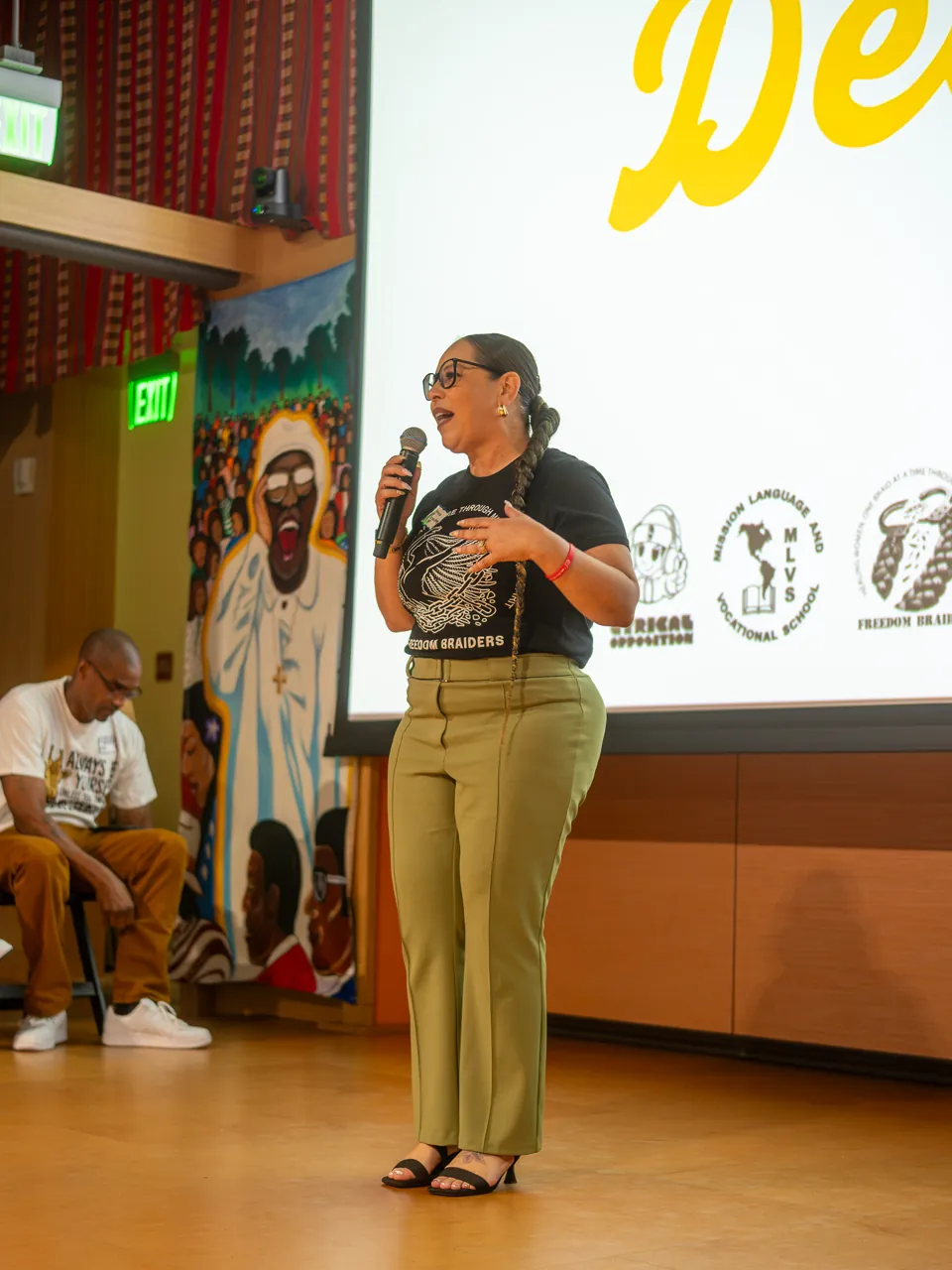 A speaker in a black shirt and olive trousers addresses guests beneath the Deville title.