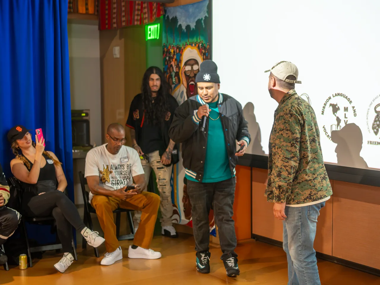 A guest in a dark jacket and green shirt joins speakers in front of the audience.