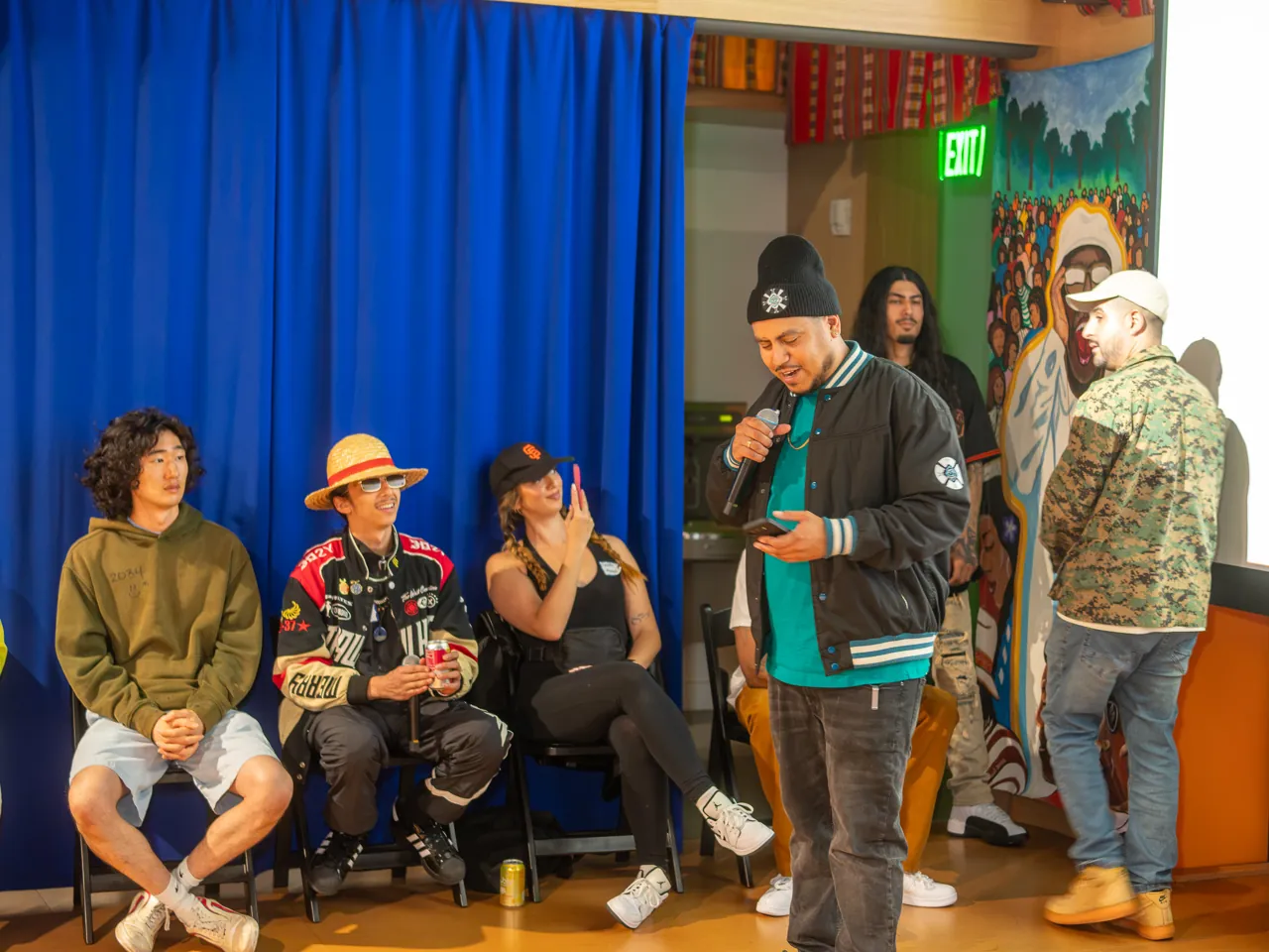 A guest in a dark jacket and green shirt joins speakers in front of the audience.