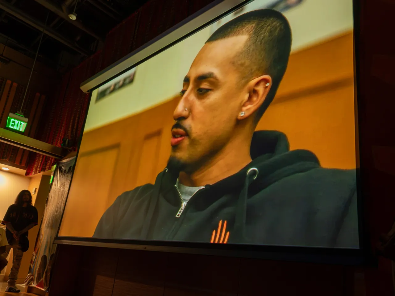 A projected interview shows a person in a dark hoodie looking downward.