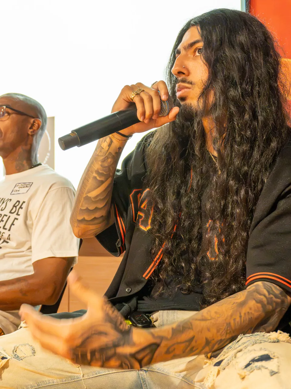 A long-haired panel participant speaks into a microphone while seated beside the screen.
