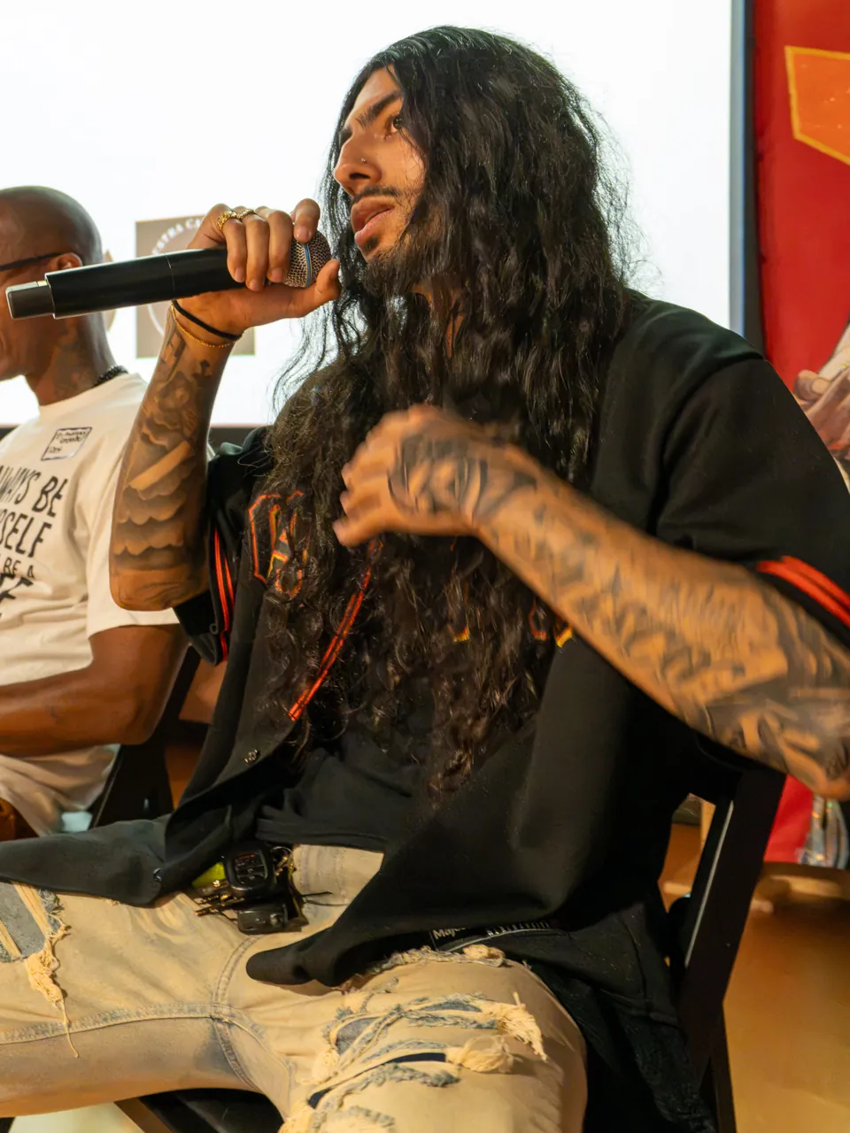 A long-haired panel participant speaks into a microphone while seated beside the screen.