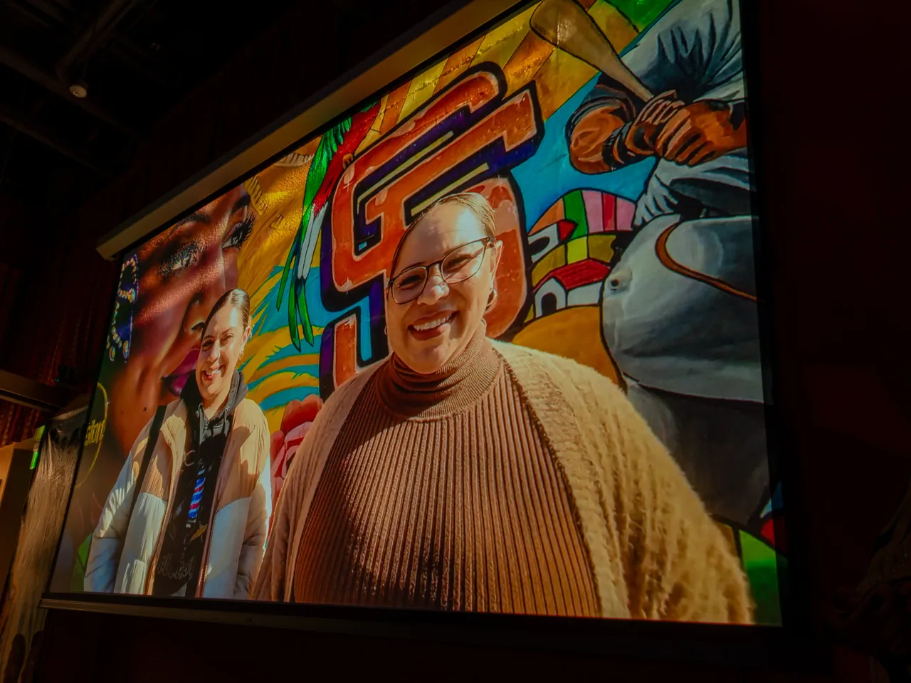 A projected interview shows a smiling person in front of a colorful mural.