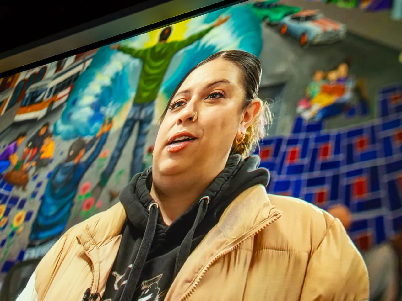 A projected interview shows a person in a tan jacket against a colorful mural.