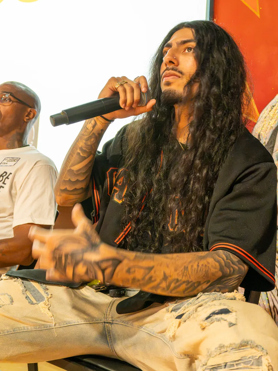A long-haired panel participant speaks into a microphone while seated beside the screen.