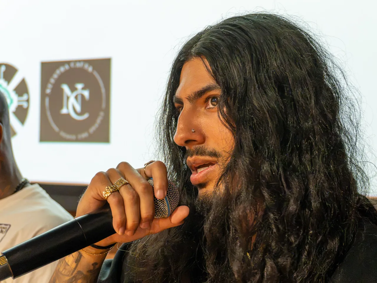 A long-haired panel participant speaks into a microphone while seated beside the screen.