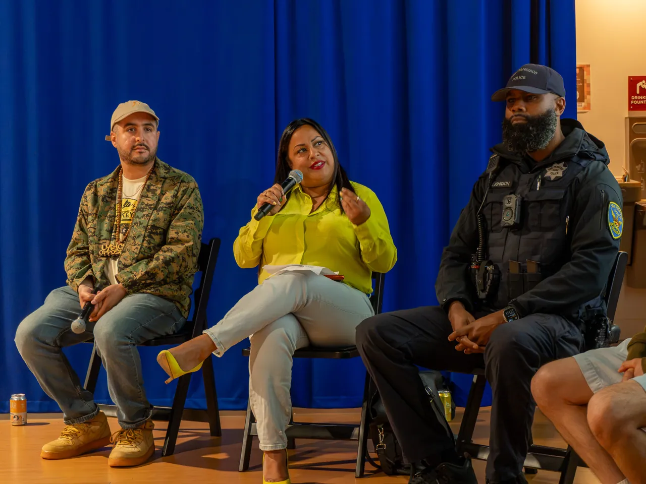 A panel participant in a bright yellow top speaks into a microphone between two seated participants.