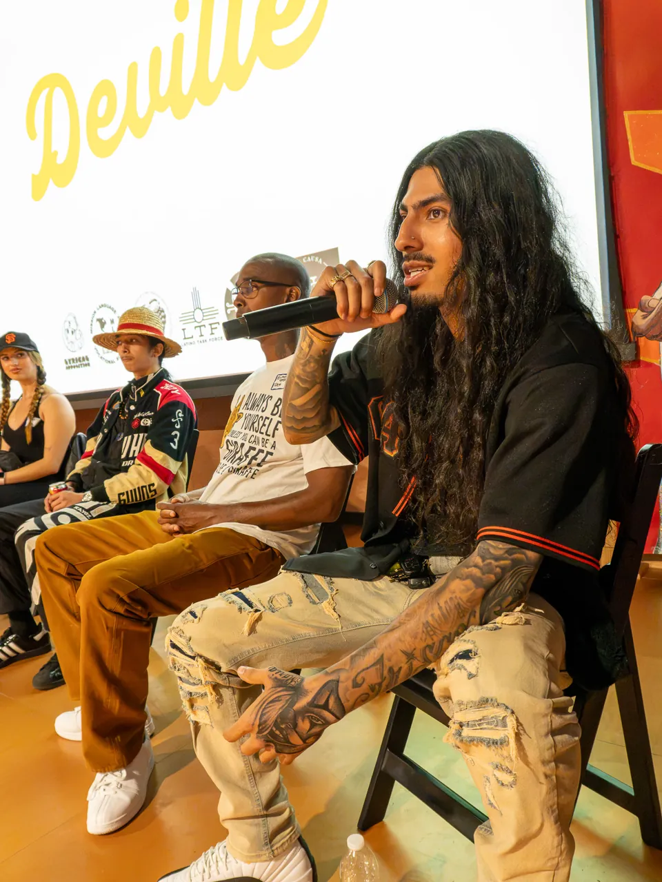 A long-haired panel participant speaks into a microphone while seated beside the screen.