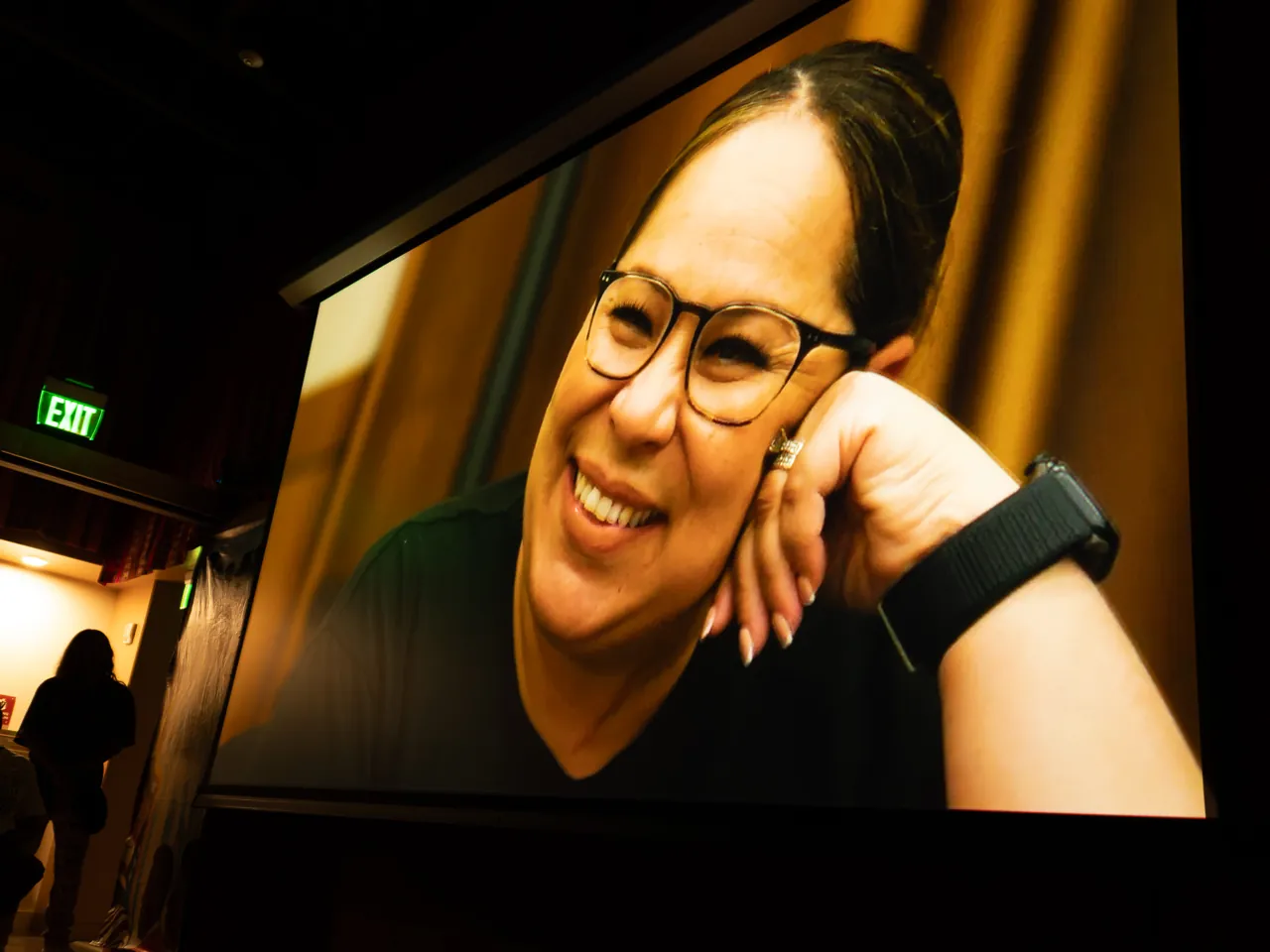 A projected close-up shows a smiling person wearing glasses.