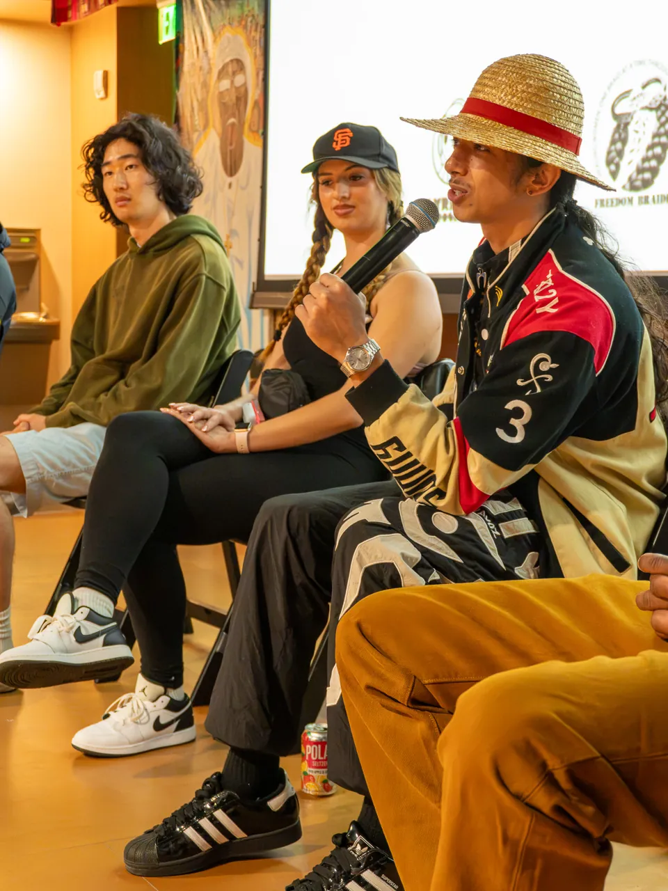 A panel participant in a straw hat speaks while two seated participants listen.