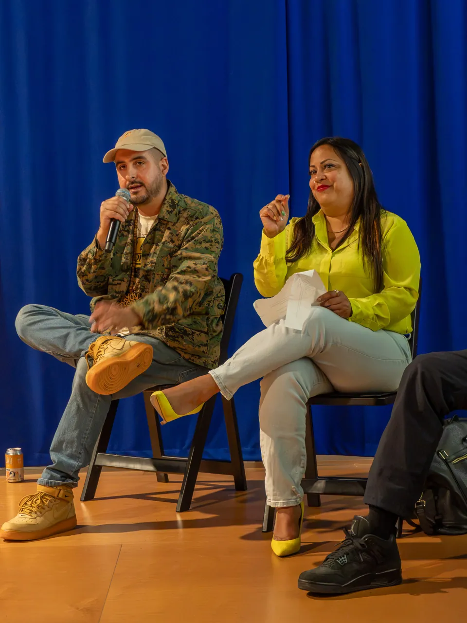 A panel participant in a patterned jacket and cap speaks beside participants in yellow and black.