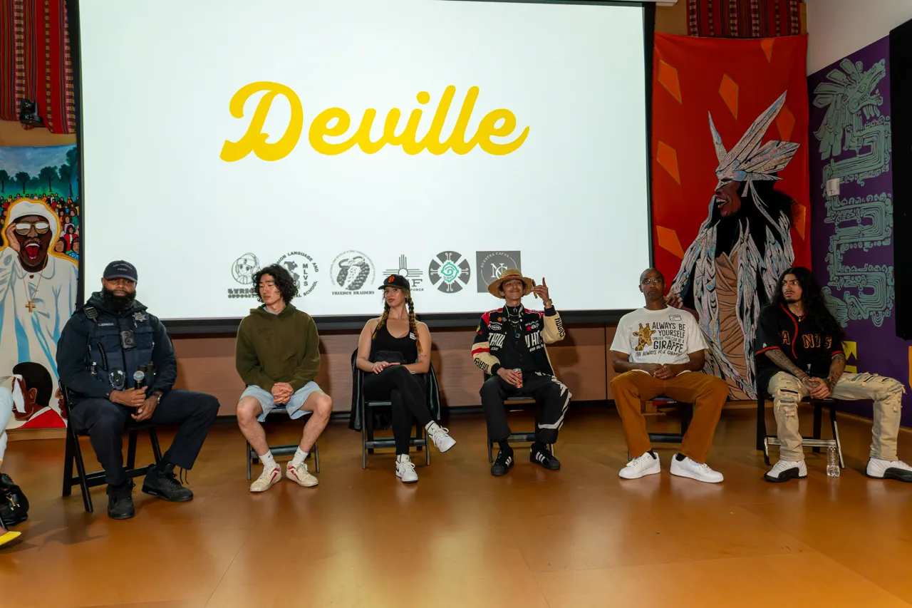Panel participants sit beneath the projected Deville title, with murals and blue curtains around them.
