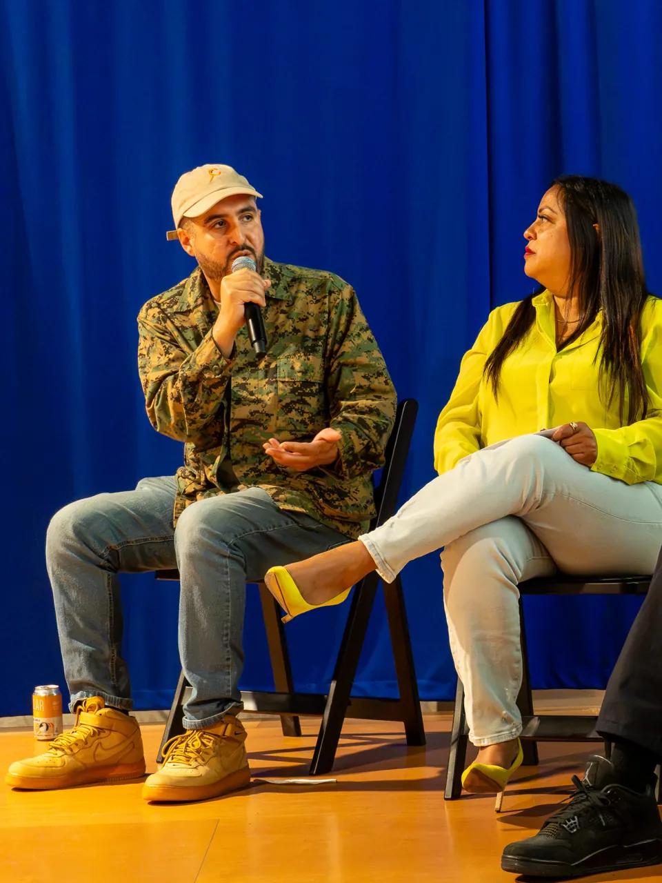 A panel participant in a patterned jacket and cap speaks beside participants in yellow and black.