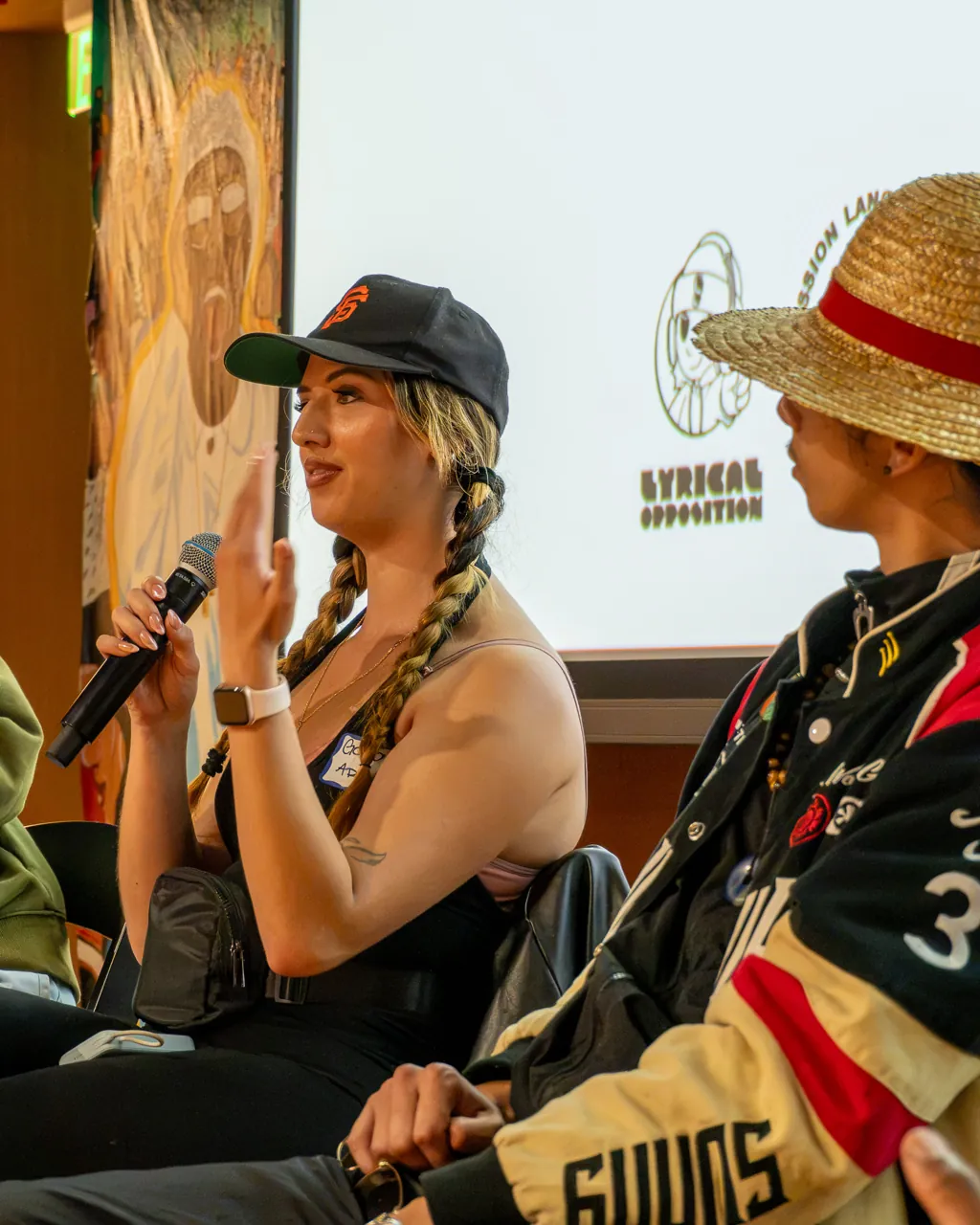 A panel participant in a black cap speaks into a microphone beside two other participants.
