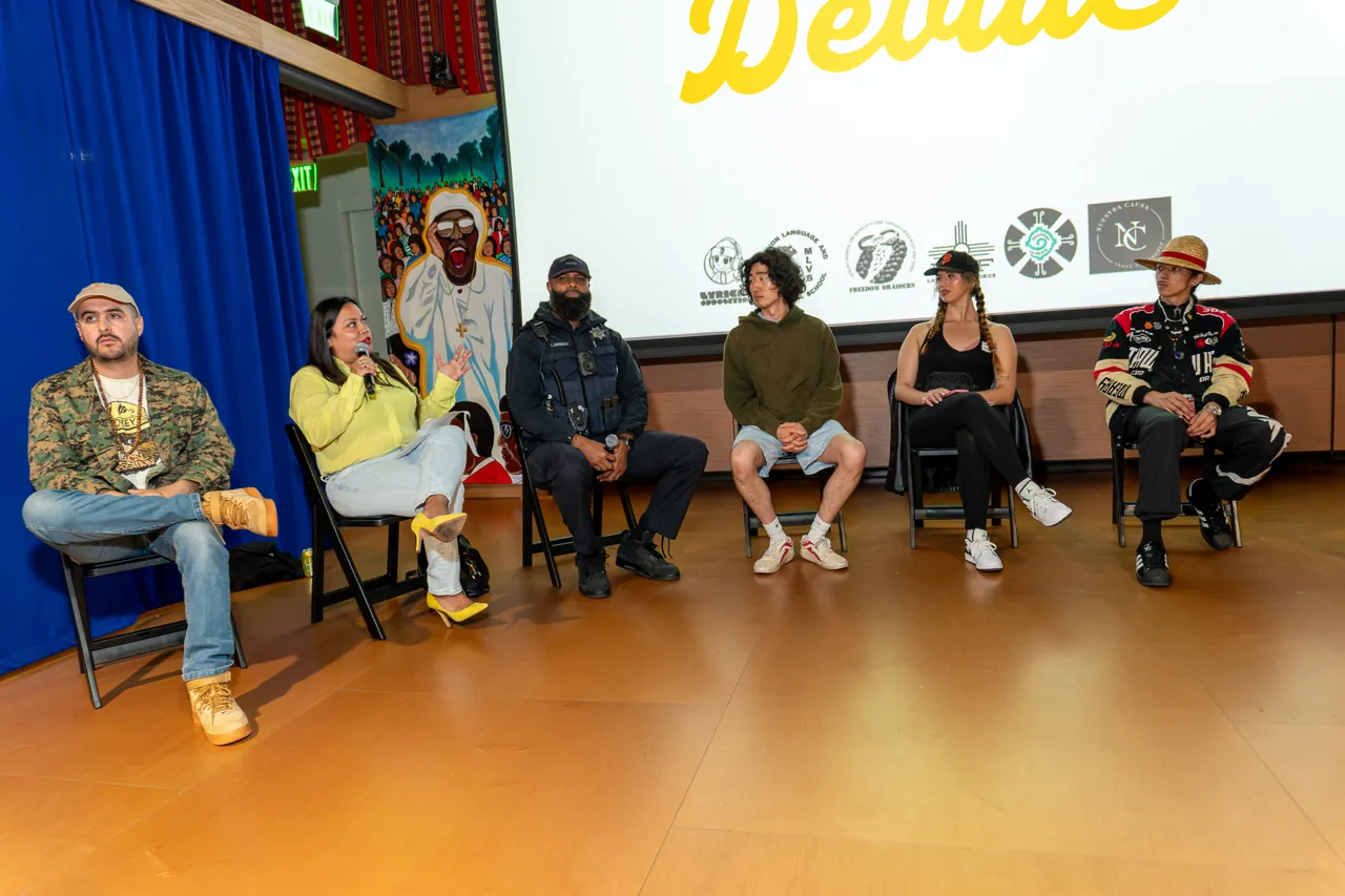 Panel participants sit beneath the projected Deville title, with murals and blue curtains around them.