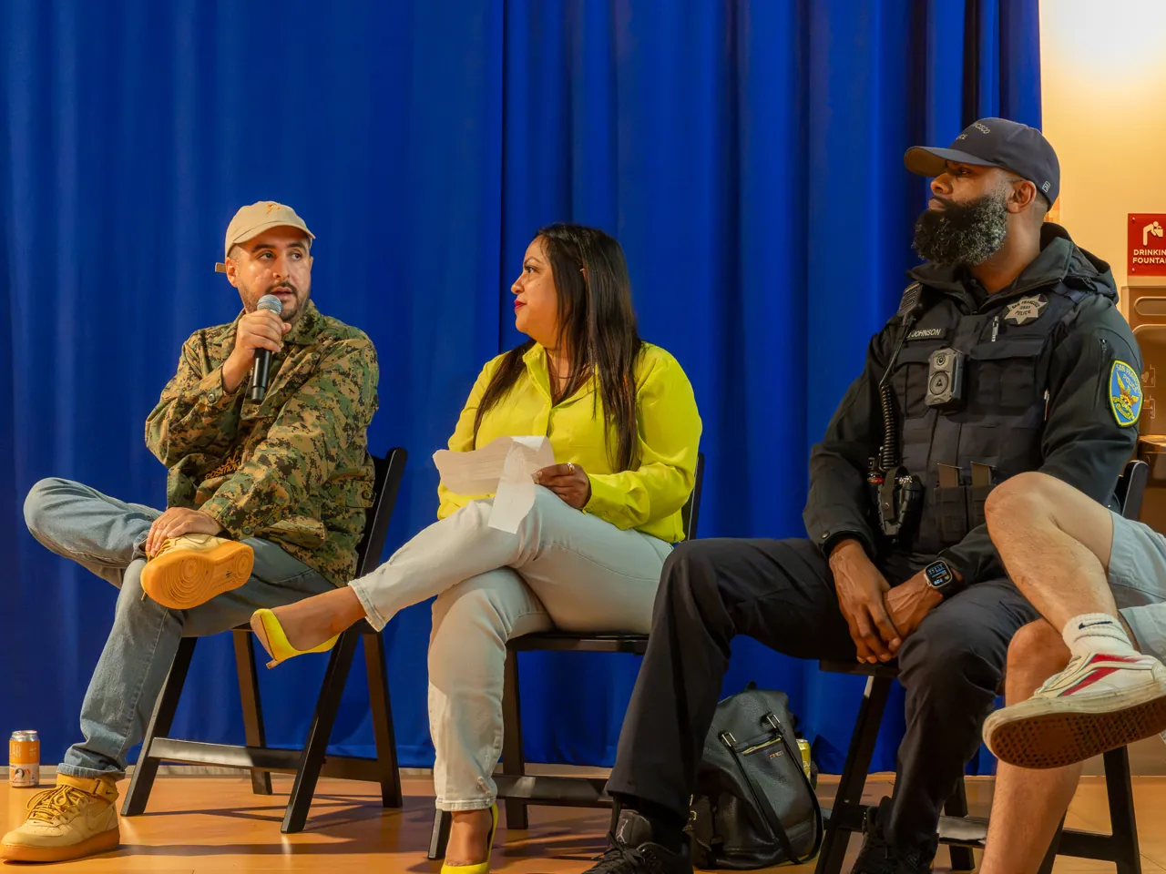 A panel participant in a patterned jacket and cap speaks beside participants in yellow and black.