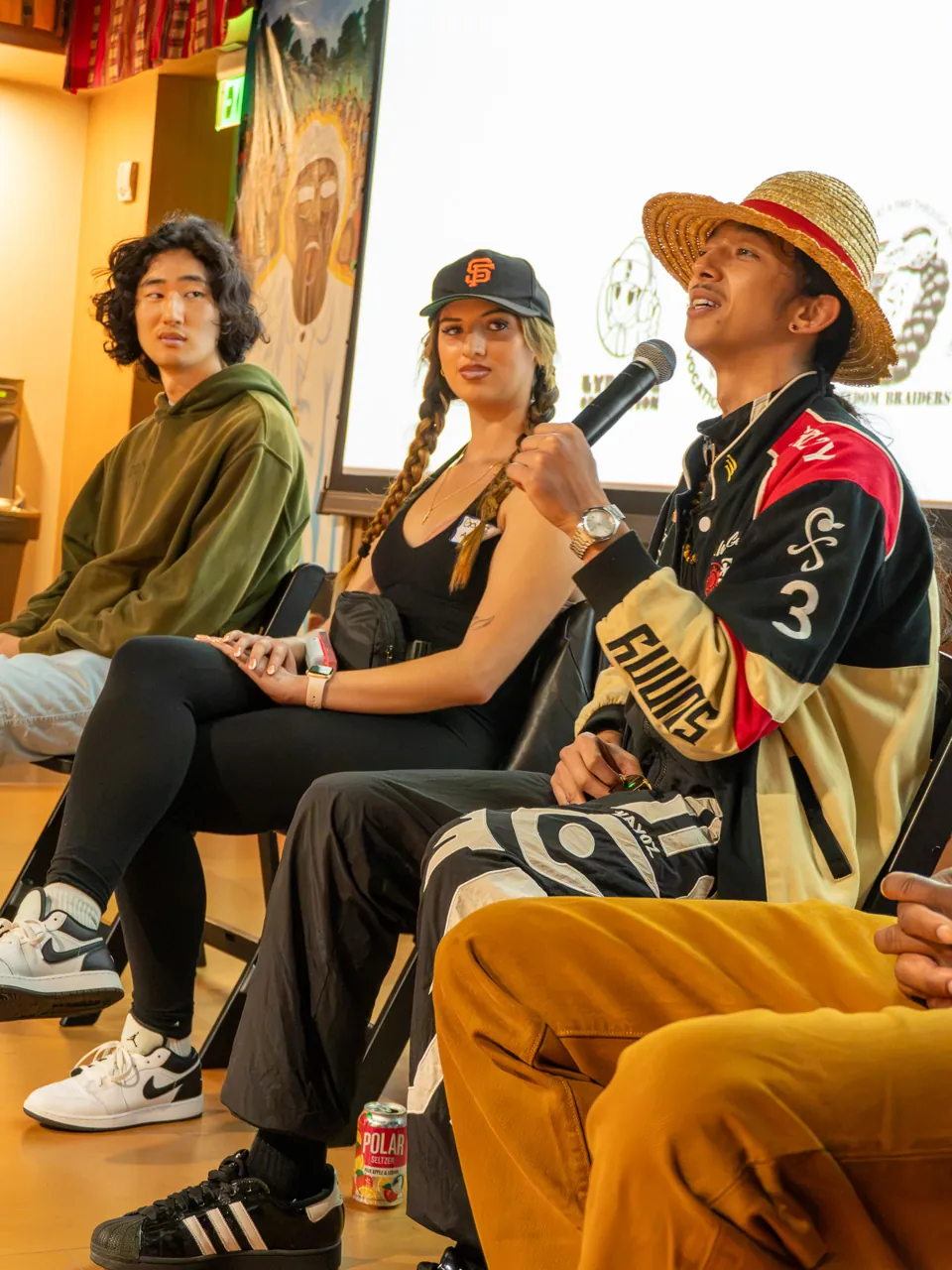 A panel participant in a straw hat speaks while two seated participants listen.