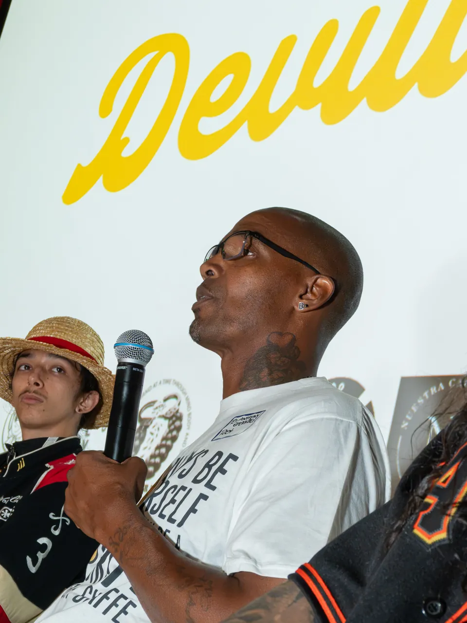 A seated panel participant in a white T-shirt speaks into a microphone.
