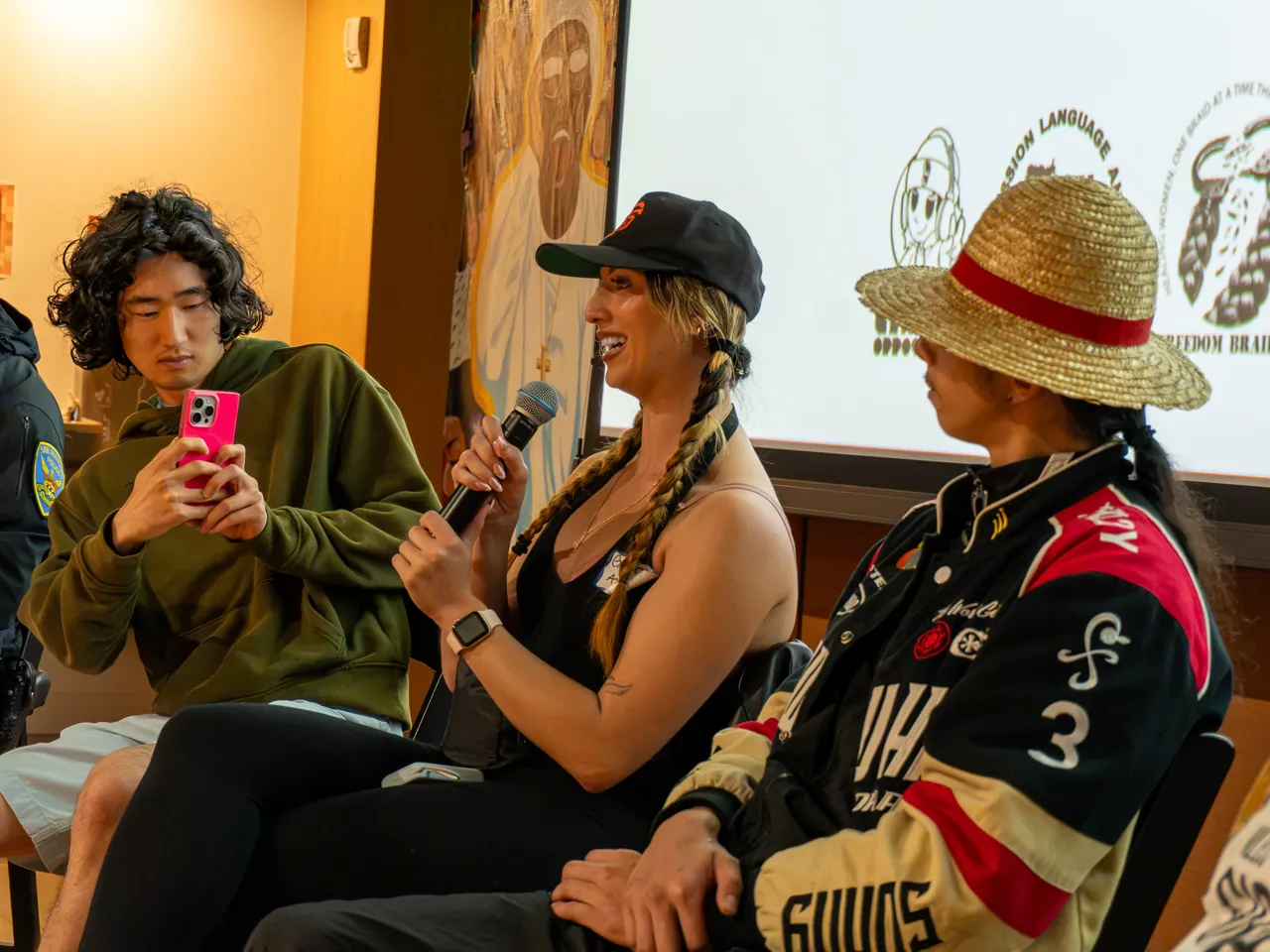 A panel participant in a black cap speaks into a microphone beside two other participants.