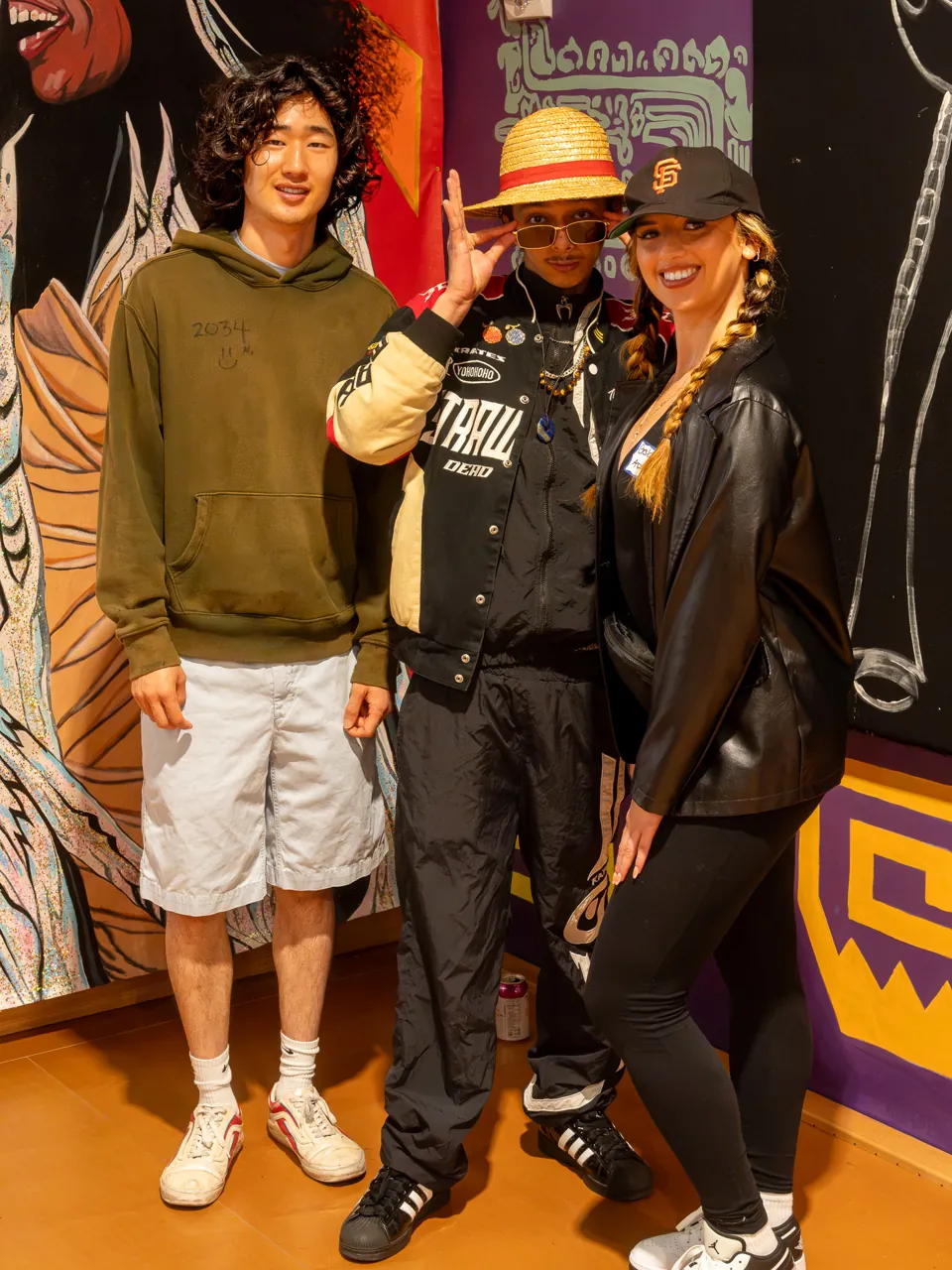 Three guests pose in front of a mural, including a person in a straw hat.