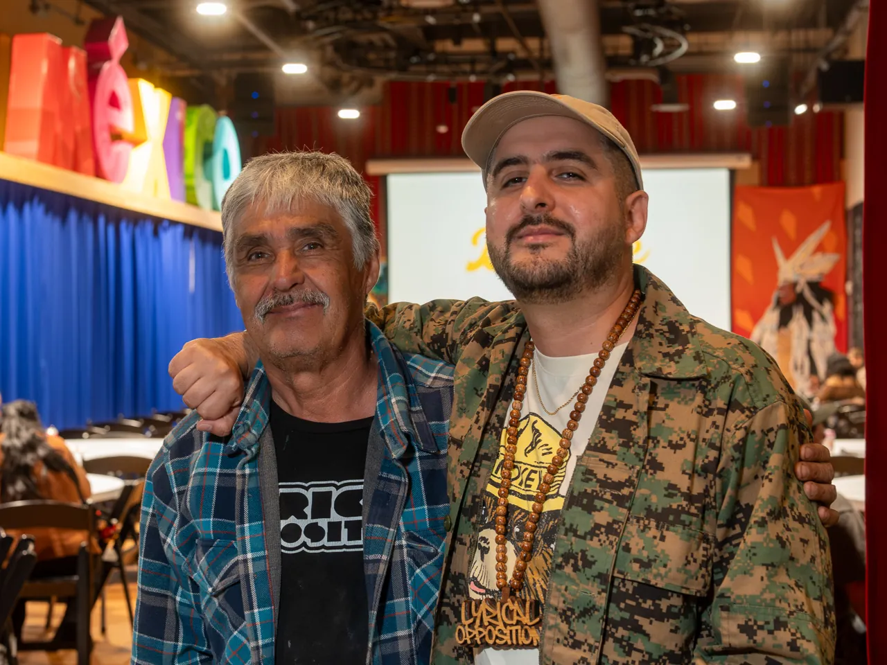 Two guests pose shoulder to shoulder, one wearing a plaid shirt and the other a patterned jacket.