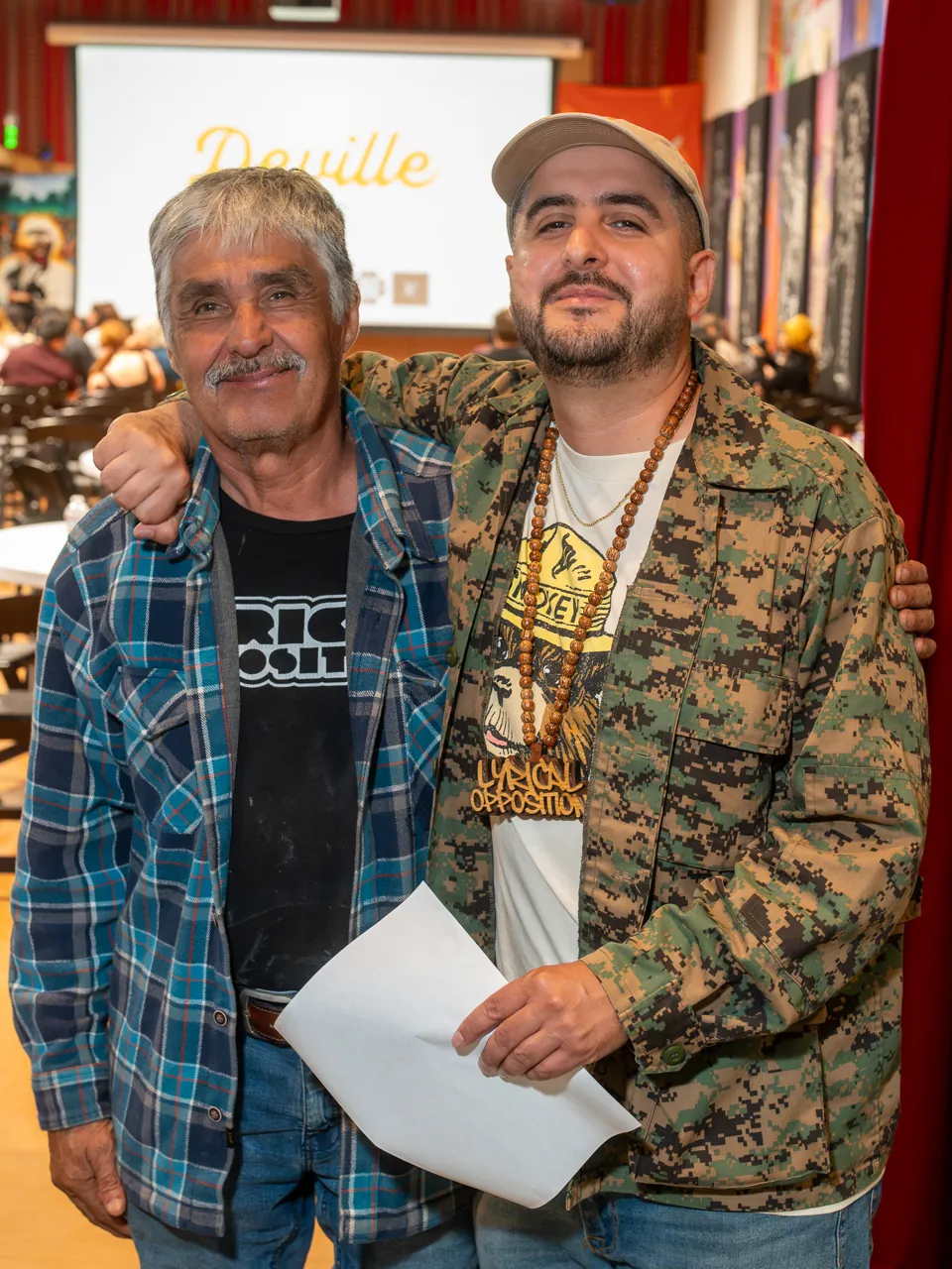Two guests pose shoulder to shoulder, one wearing a plaid shirt and the other a patterned jacket.