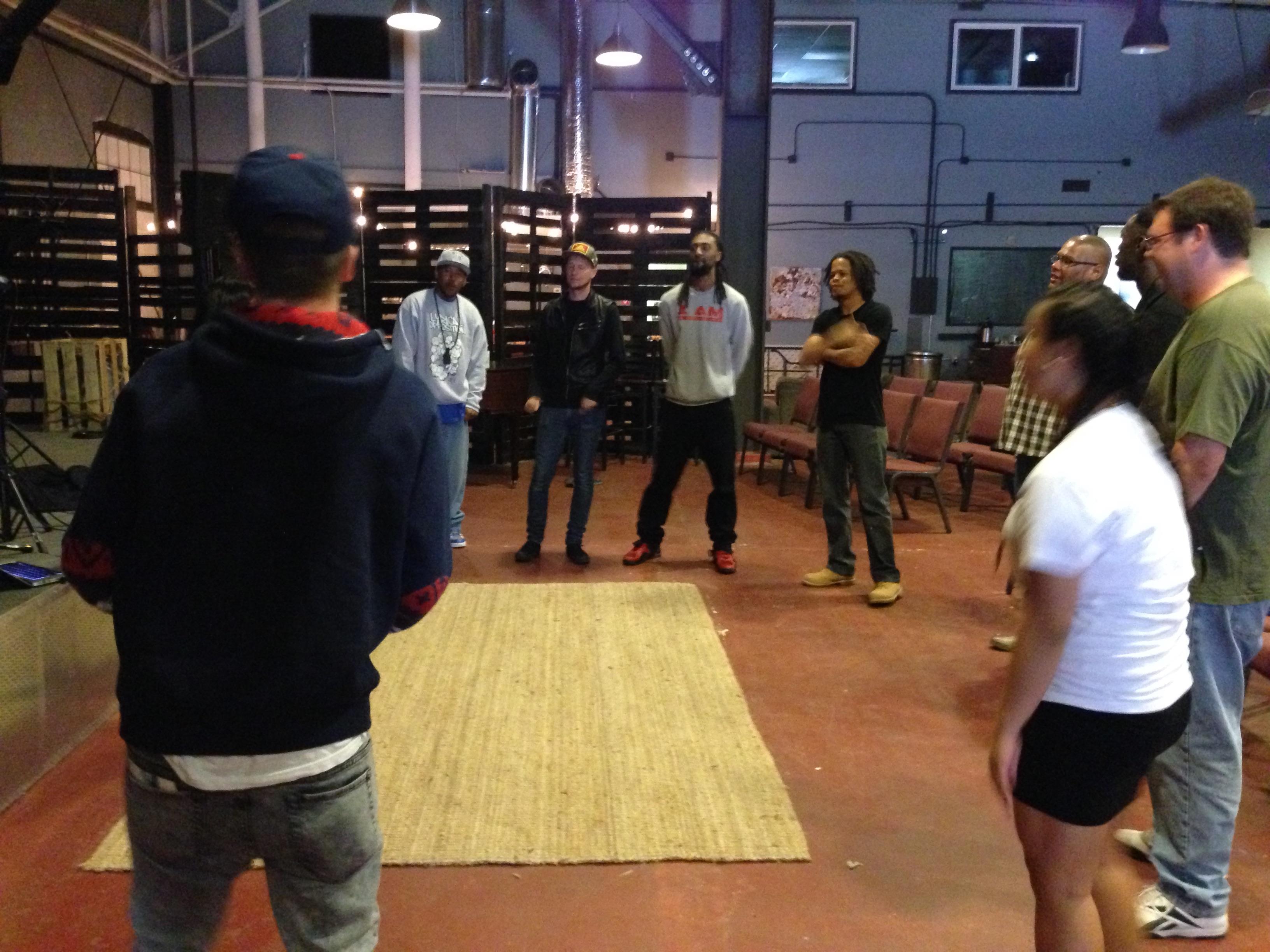 A group of people standing in a circle around a woven rug in a large room with string lights and wooden pallet walls.
