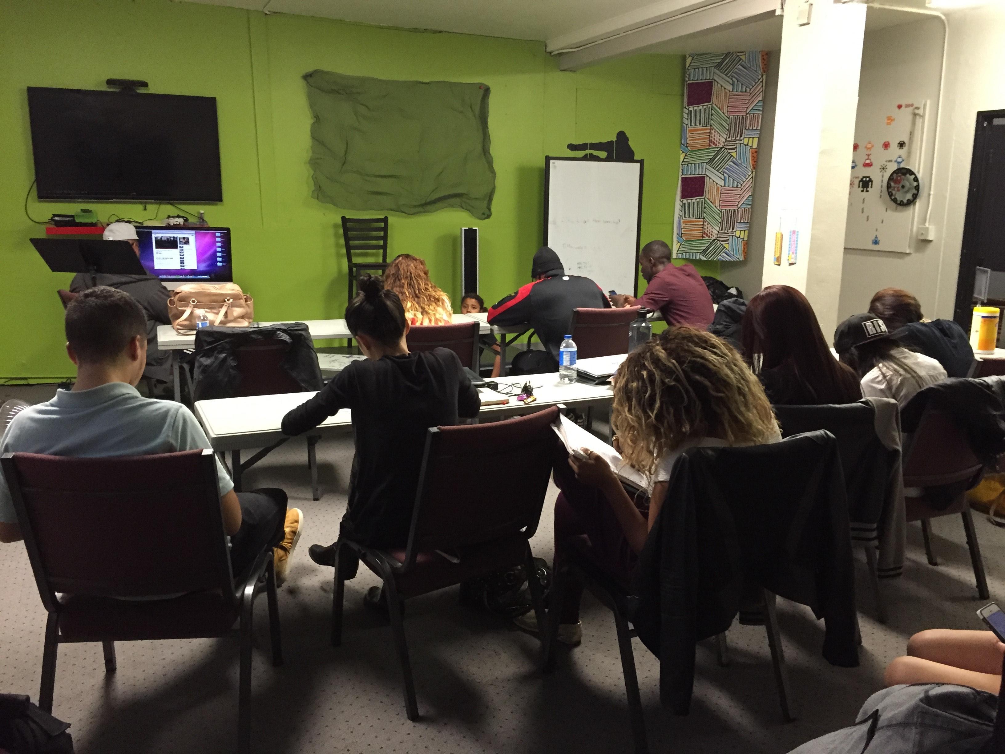 People seated at tables and on chairs writing in notebooks in a room with green walls, a whiteboard, and a wall mounted screen.