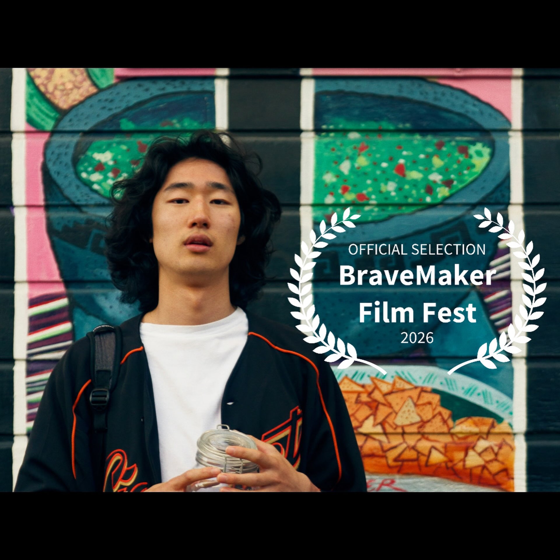 A young man with long dark hair in a black jacket over a white T-shirt holds a glass jar in front of a painted mural of food bowls and tortilla chips, beside white laurels reading Official Selection, BraveMaker Film Fest 2026.