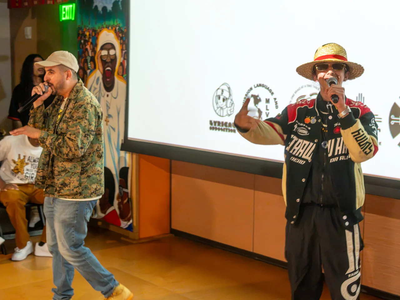 Two performers hold microphones in front of a projection screen and mural at the Deville community screening.
