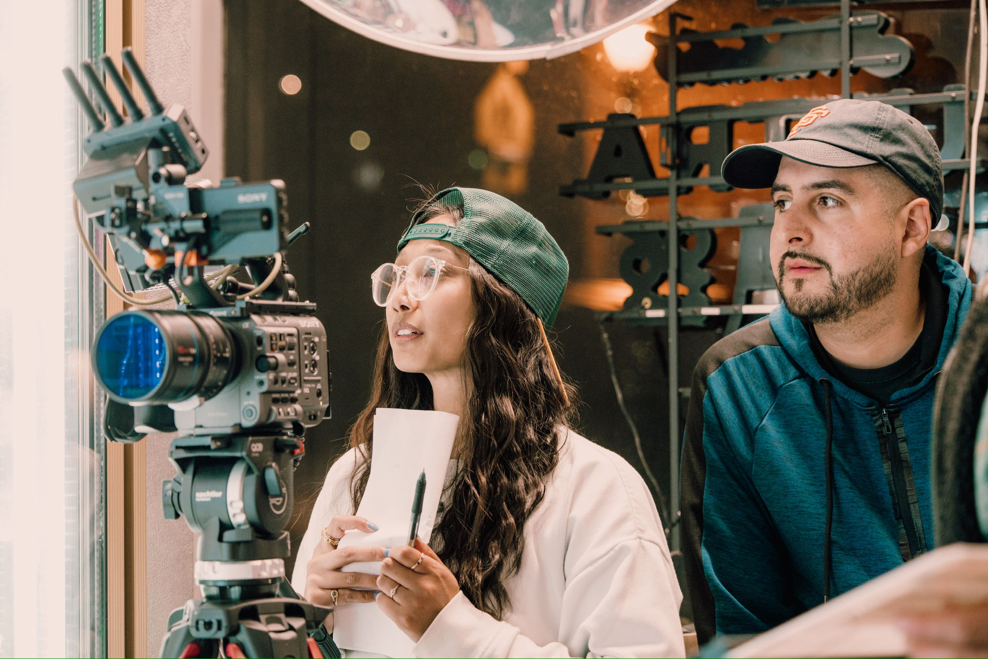 Two crew members beside a film camera outside a storefront, one holding production notes.