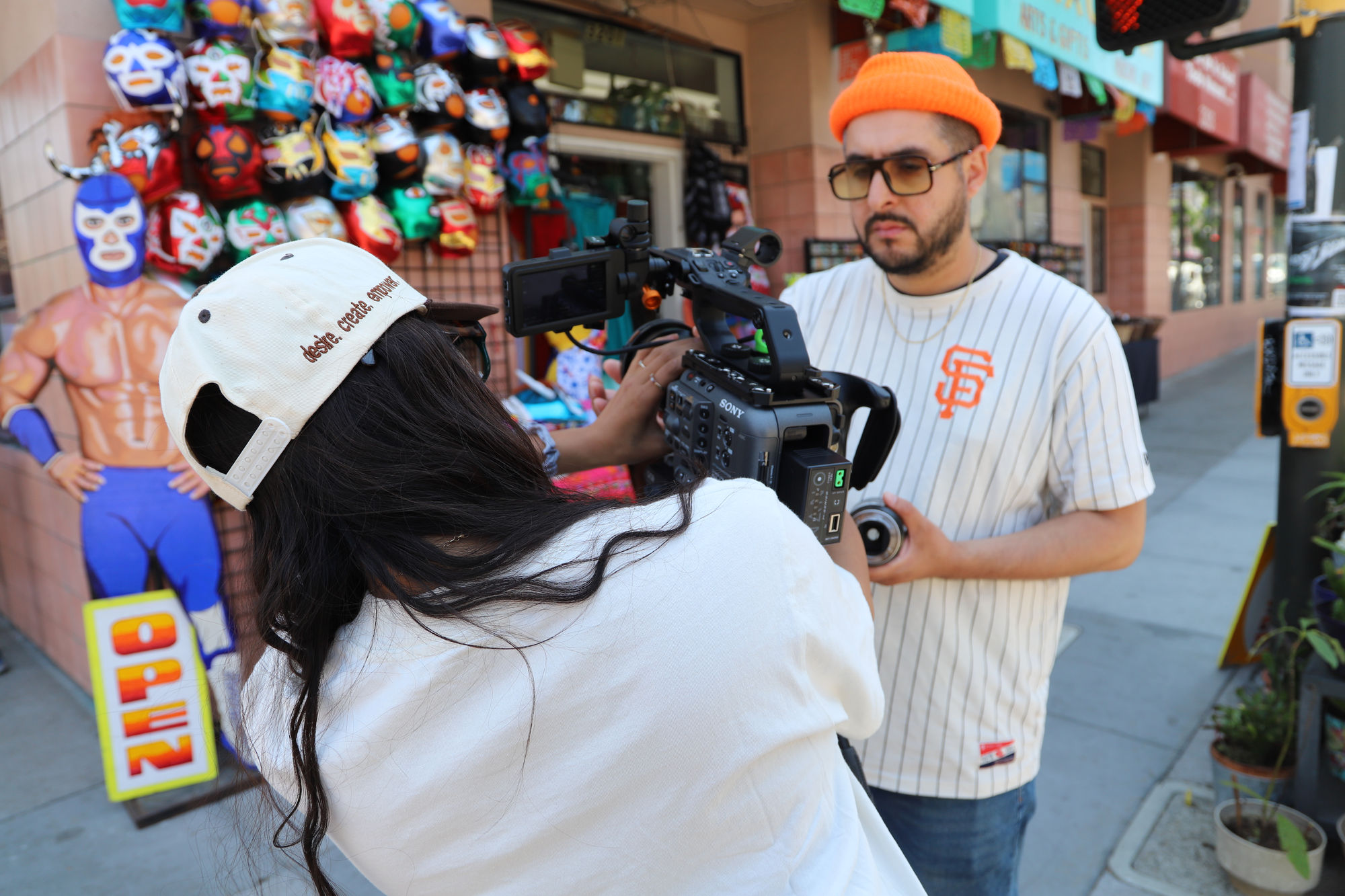A camera operator films on a sidewalk outside a shop hung with lucha libre masks.