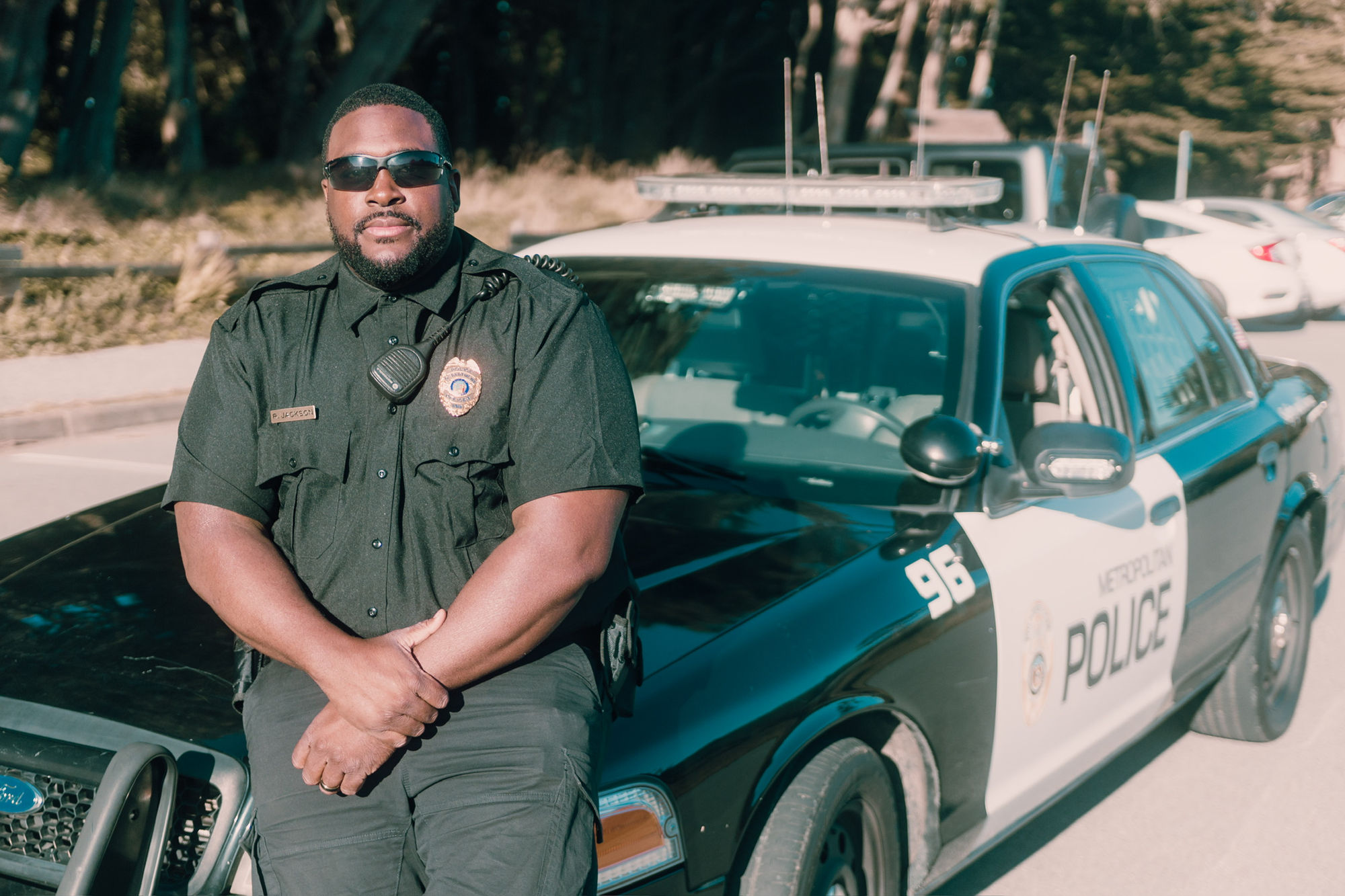 An actor in uniform and sunglasses leaning against a police cruiser.