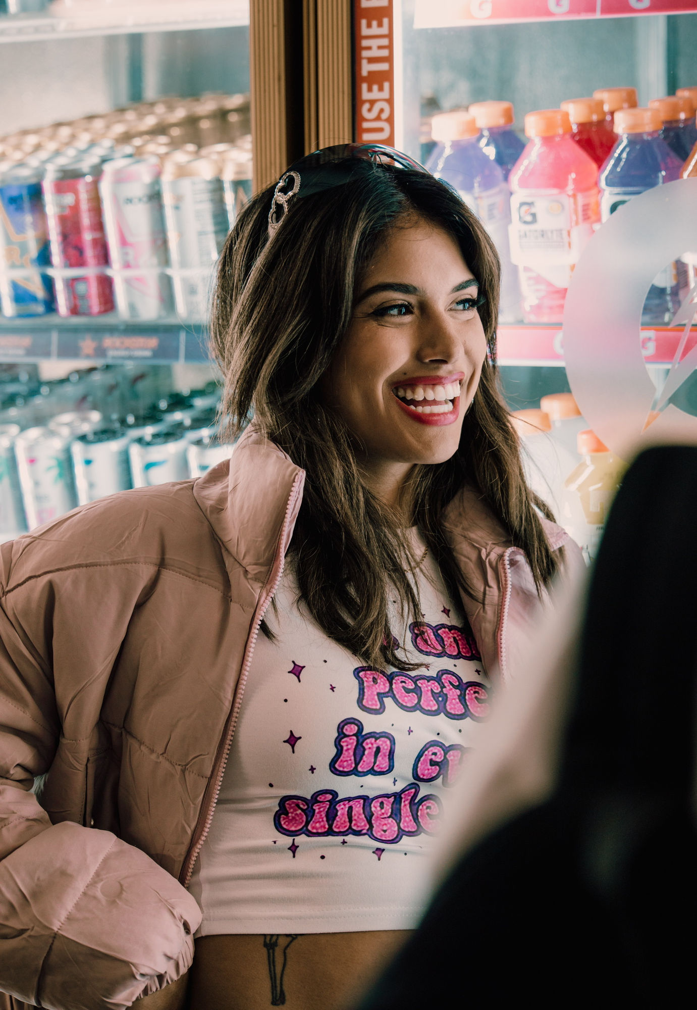 A young woman in a pink jacket laughing beside the drinks cooler in a corner store.