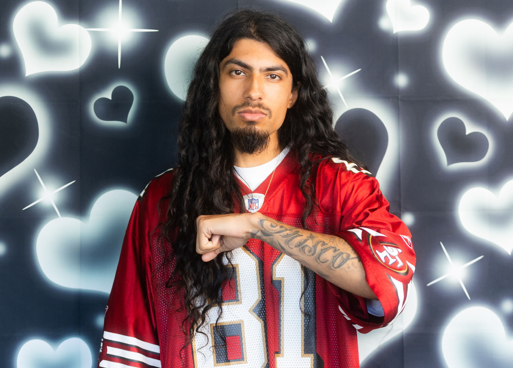 An actor in a red 49ers jersey posing against an airbrushed heart backdrop.