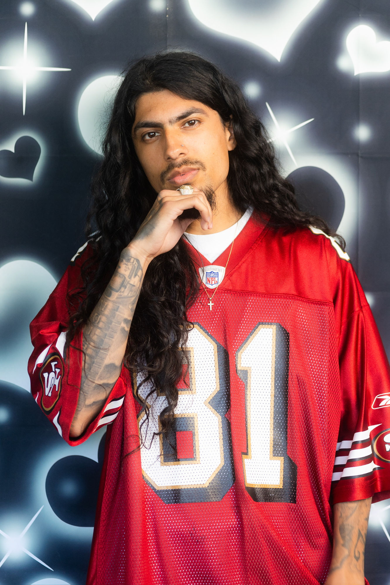An actor in a red 49ers jersey, hand to his chin, against an airbrushed heart backdrop.