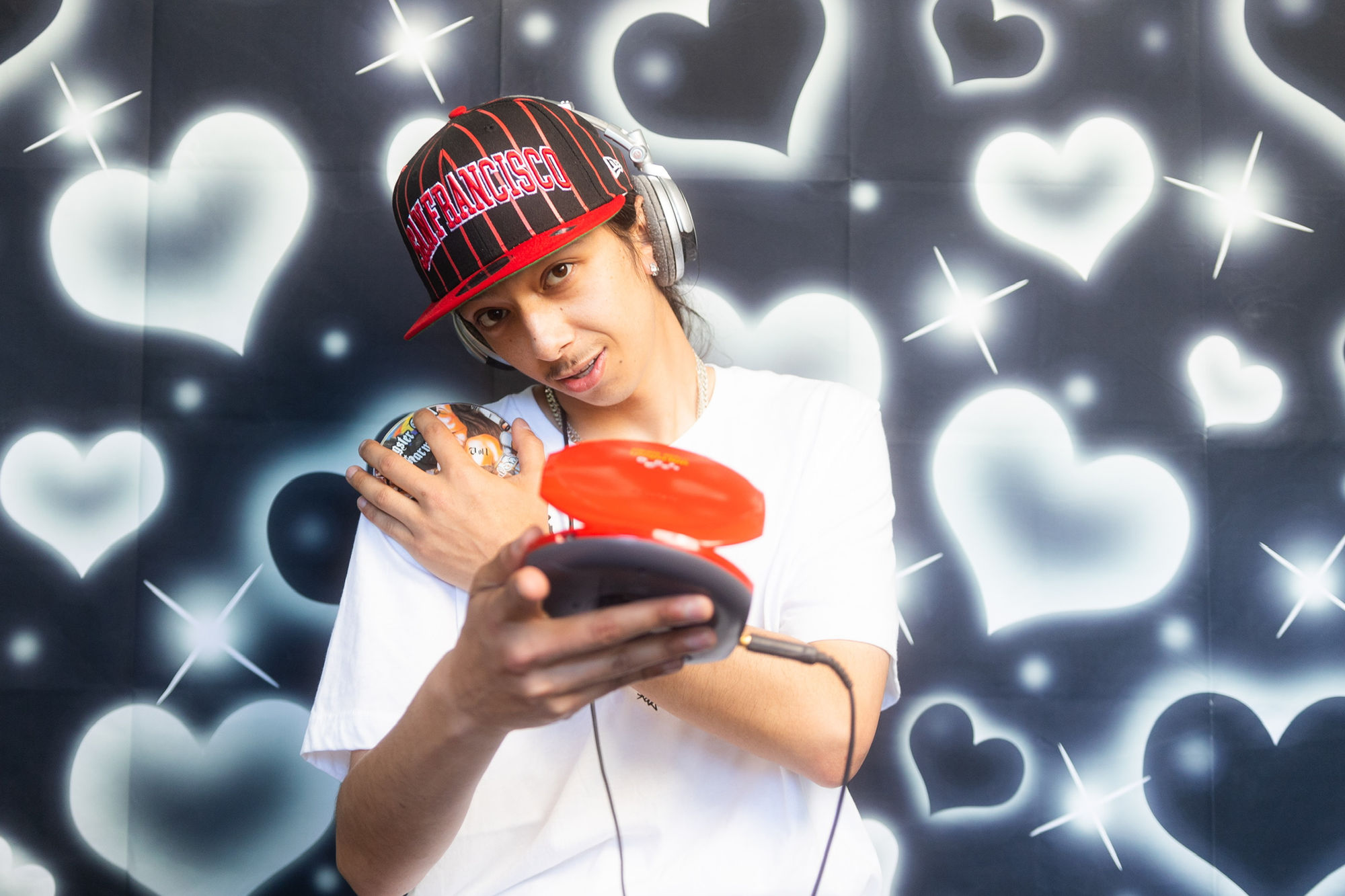 A performer in a red cap holding an open portable CD player against a heart backdrop.