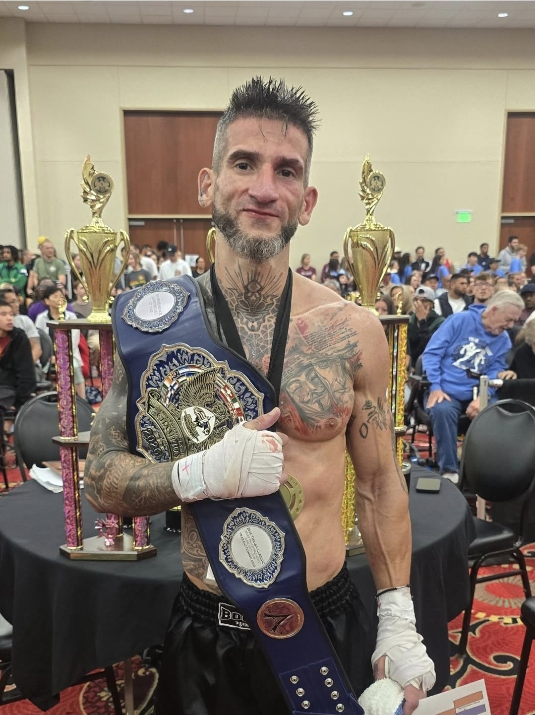Tattooed male boxer holding championship belt, standing in front of large trophies and crowd in event hall.