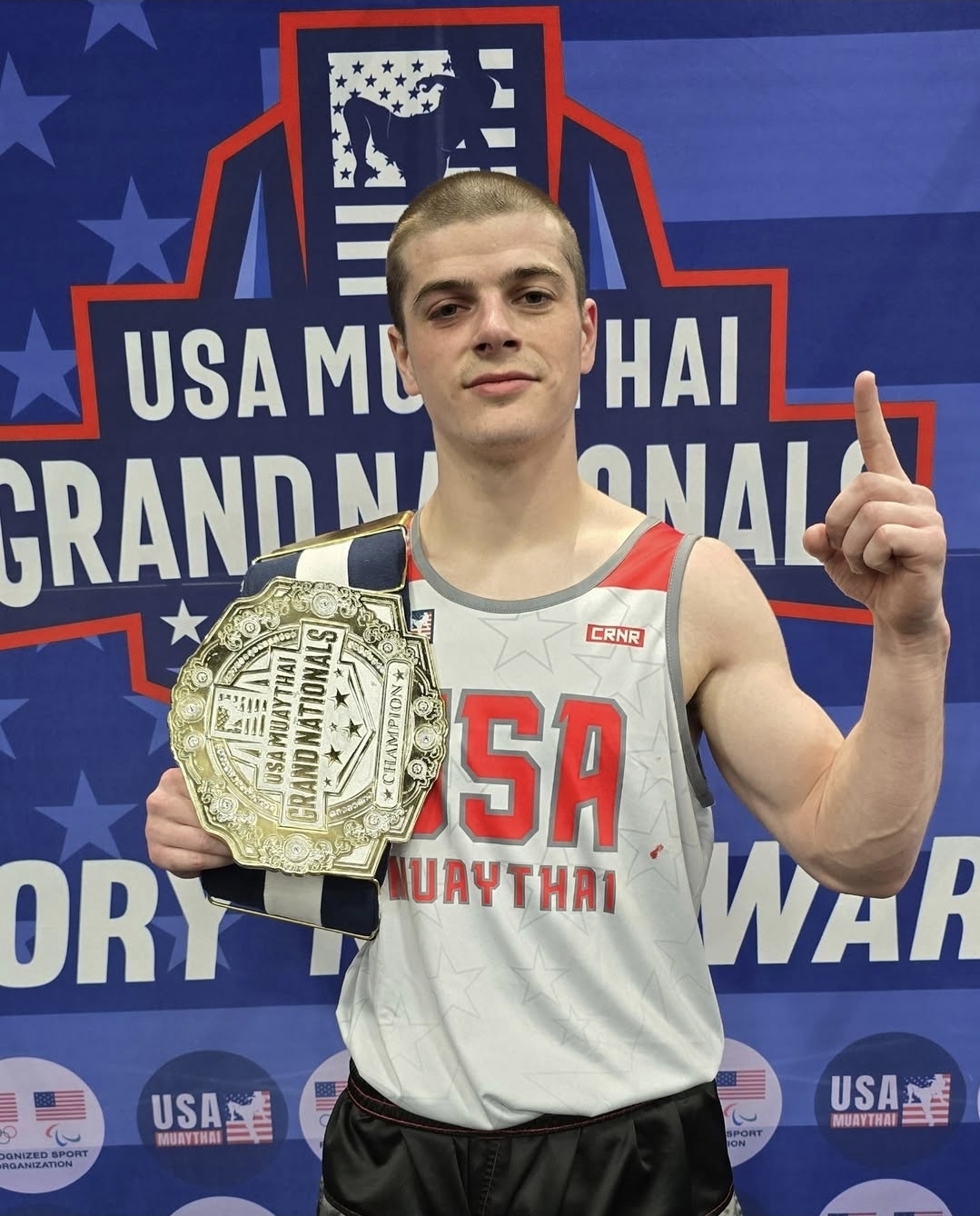 Male athlete holding a USA Muay Thai Grand Nationals champion belt and raising one finger.