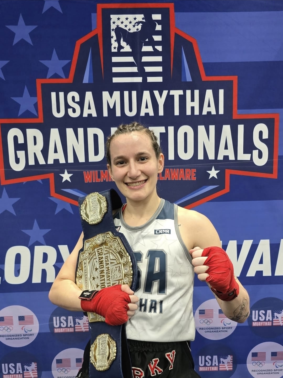 Smiling female Muay Thai champion posing with a championship belt at USA Muay Thai Grand Nationals.