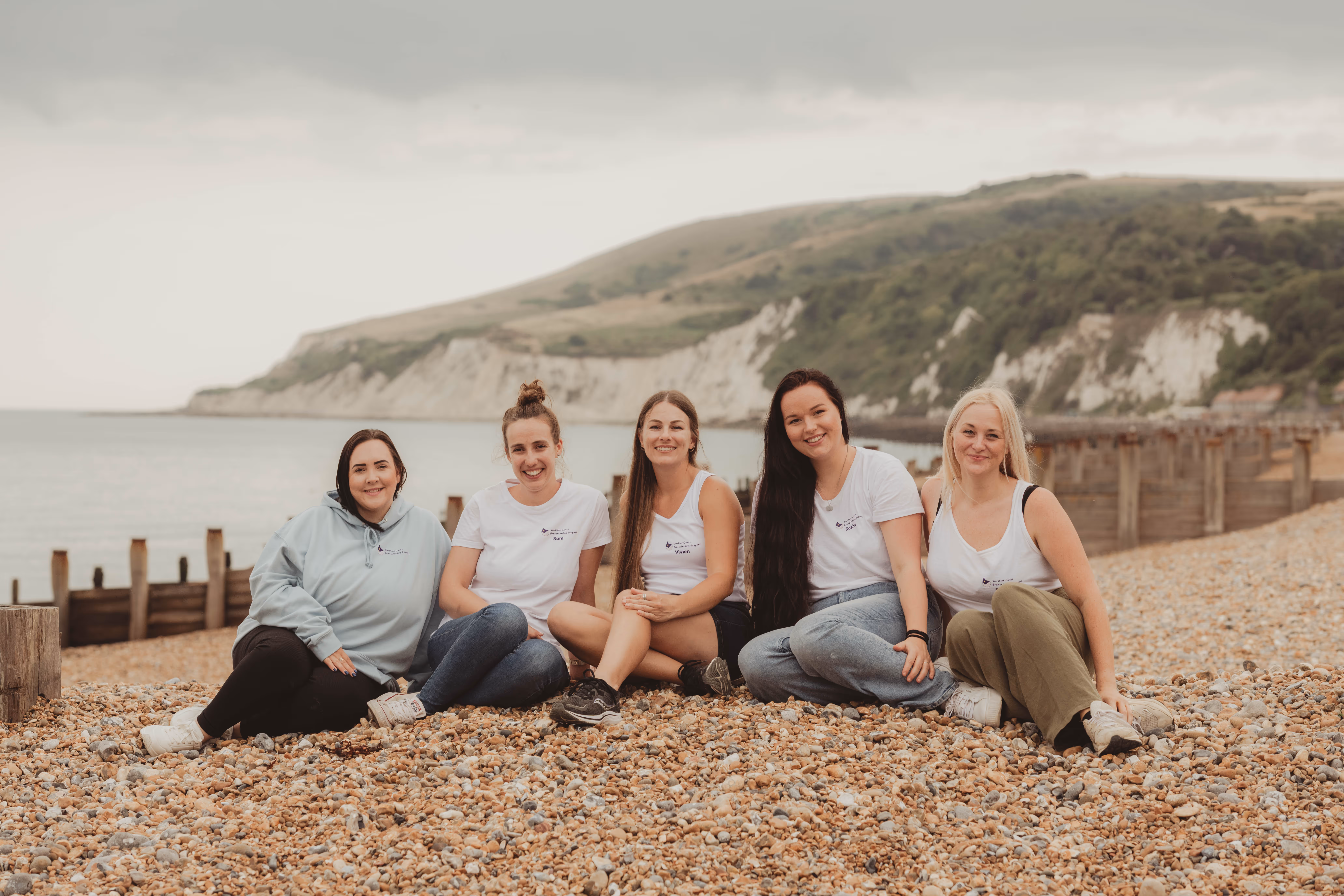 Five people sitting on a pebble beach, smiling and wearing casual clothing.